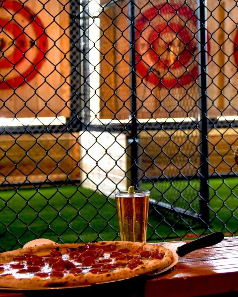 A slice of pepperoni pizza, a glass of beer with a lime wedge, on a wooden table with a chain-link fence in the background.