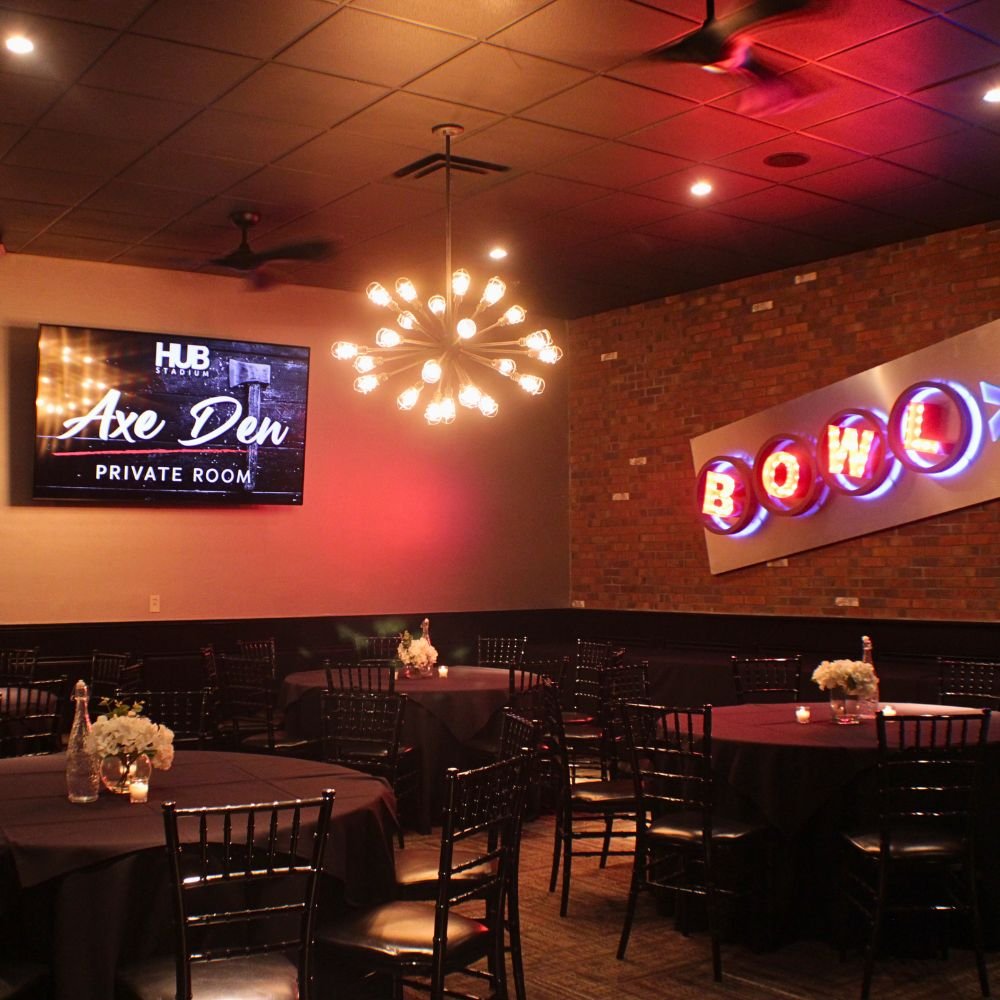 Interior of a private room at HUB Stadium called Axe Den, featuring dim lighting, round tables with dark tablecloths, decorative centerpieces, and signs reading 'BOWL' and 'Axe Den' on the wall.