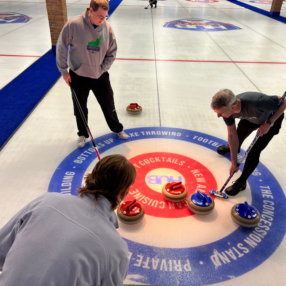 Four people are playing curling on an indoor rink. The players are positioned around the target area, called the house, which has red and blue stones. One person is delivering a red stone, while others are observing or preparing to deliver stones.