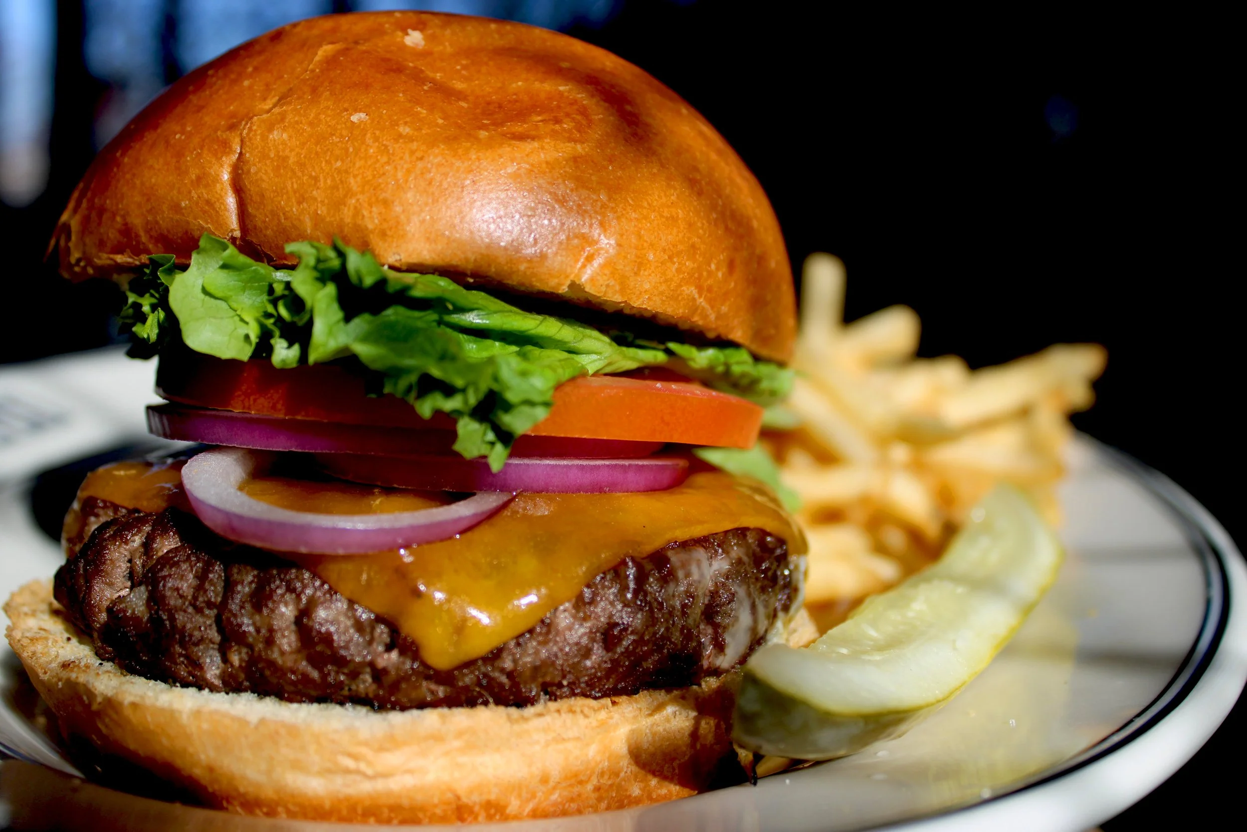 Close-up of a cheeseburger with lettuce, tomato, red onion, melted cheese, and beef patty on a bun, served with French fries and pickles on a plate.