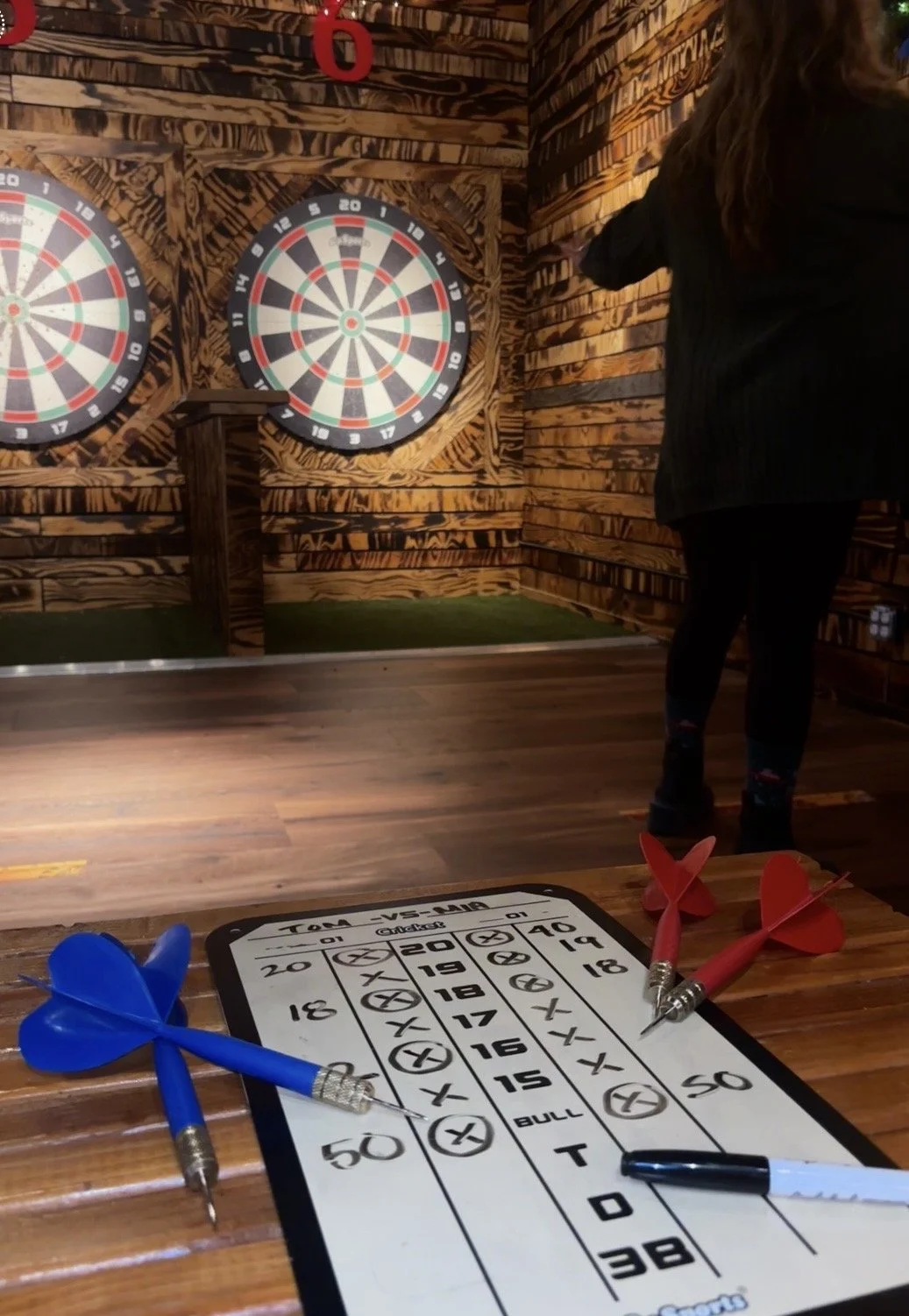 Dart game setup with a score sheet and darts, two blue and two red, on a wooden table in a room with two electronic dartboards mounted on a wooden wall in the background.