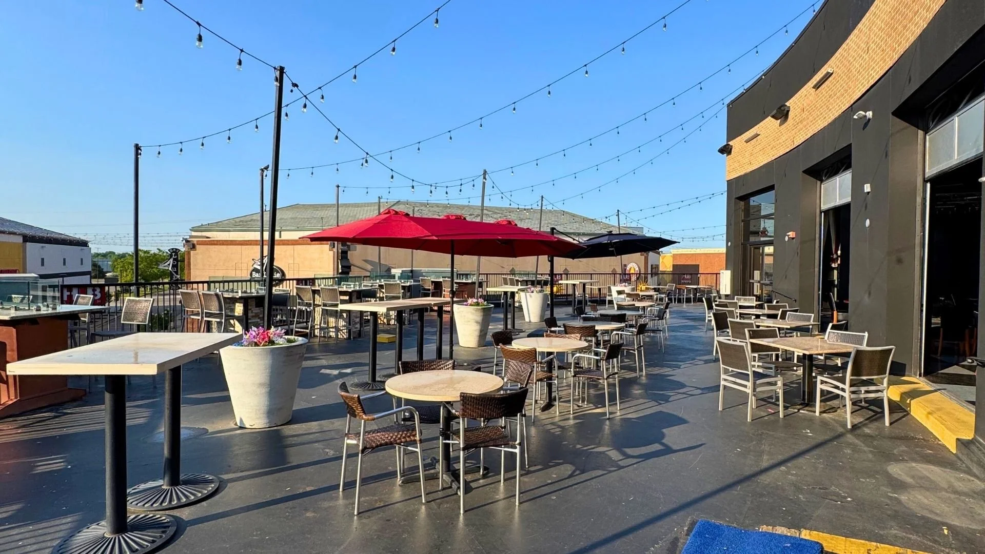 An outdoor rooftop dining area with tables, chairs, and large red and black umbrellas, decorated with string lights against a clear blue sky.