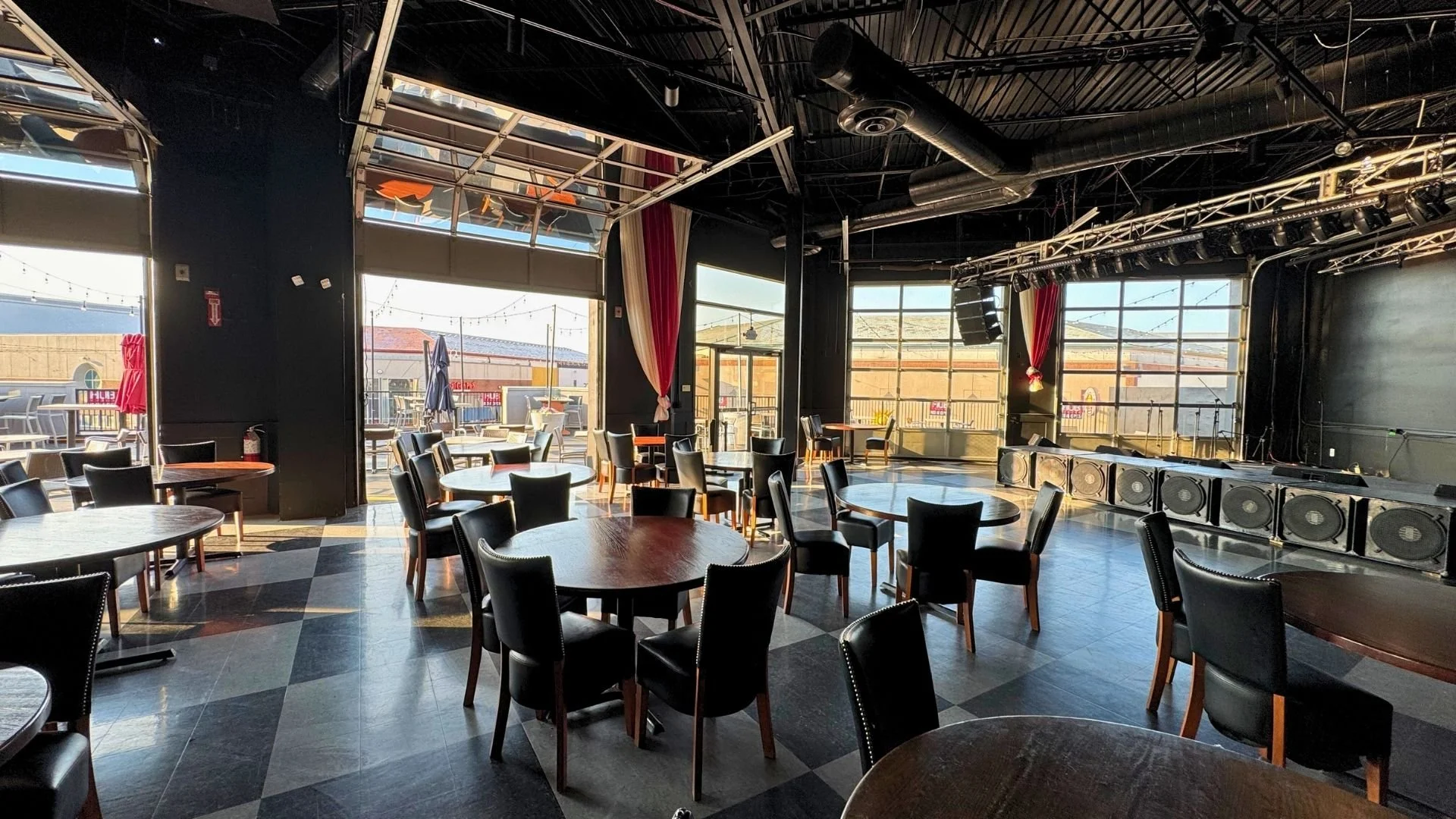 Empty indoor stage area with black flooring and multiple round tables with chairs, large windows showing outdoor patio area with tables and umbrellas, stage equipped with large speakers, lighting, and sound equipment, and ceiling with black-painted d
