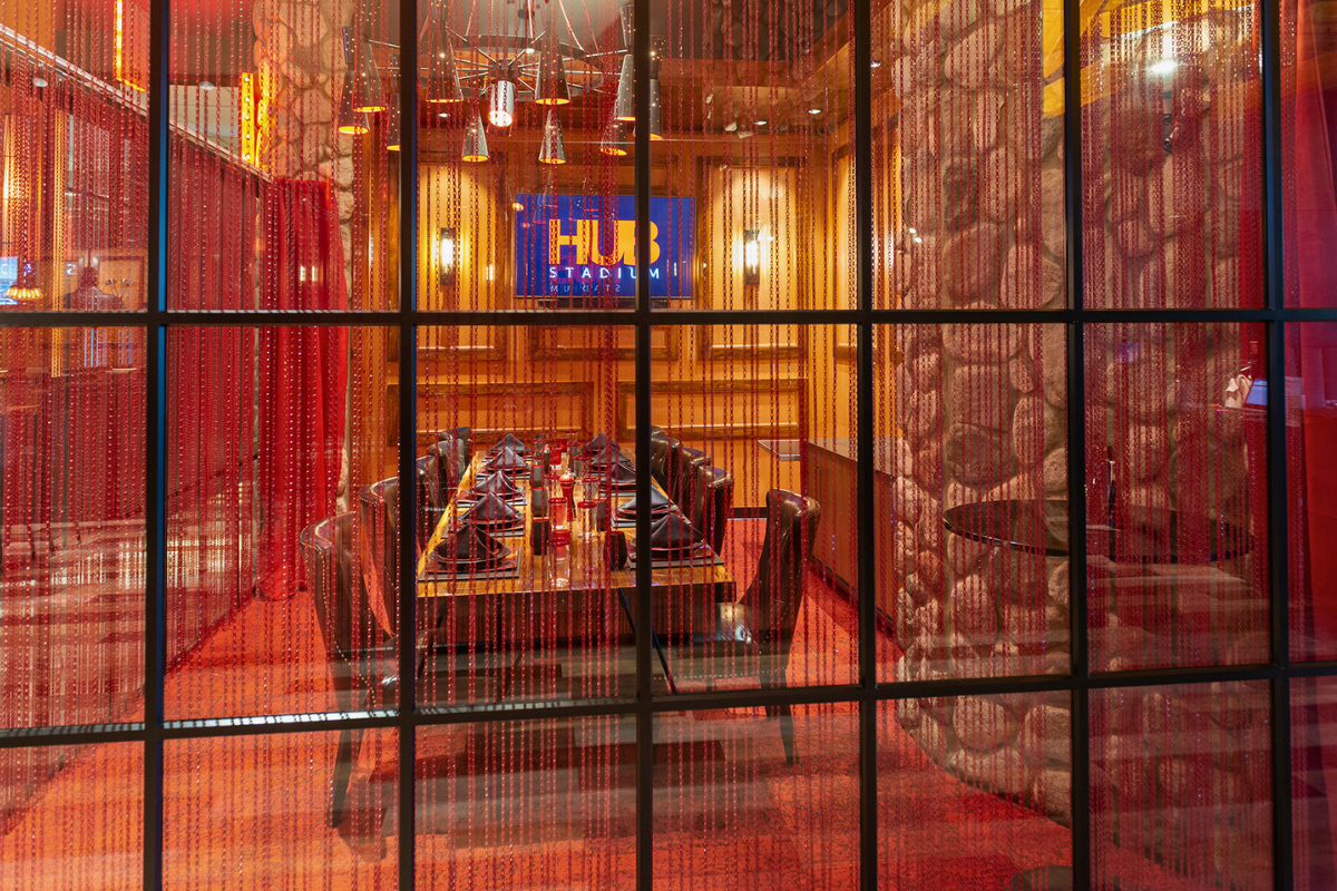 Interior view of a restaurant's private dining area with a long table, red curtains, decorative lights, and a branded TV screen displaying 'HUB Stadium.'