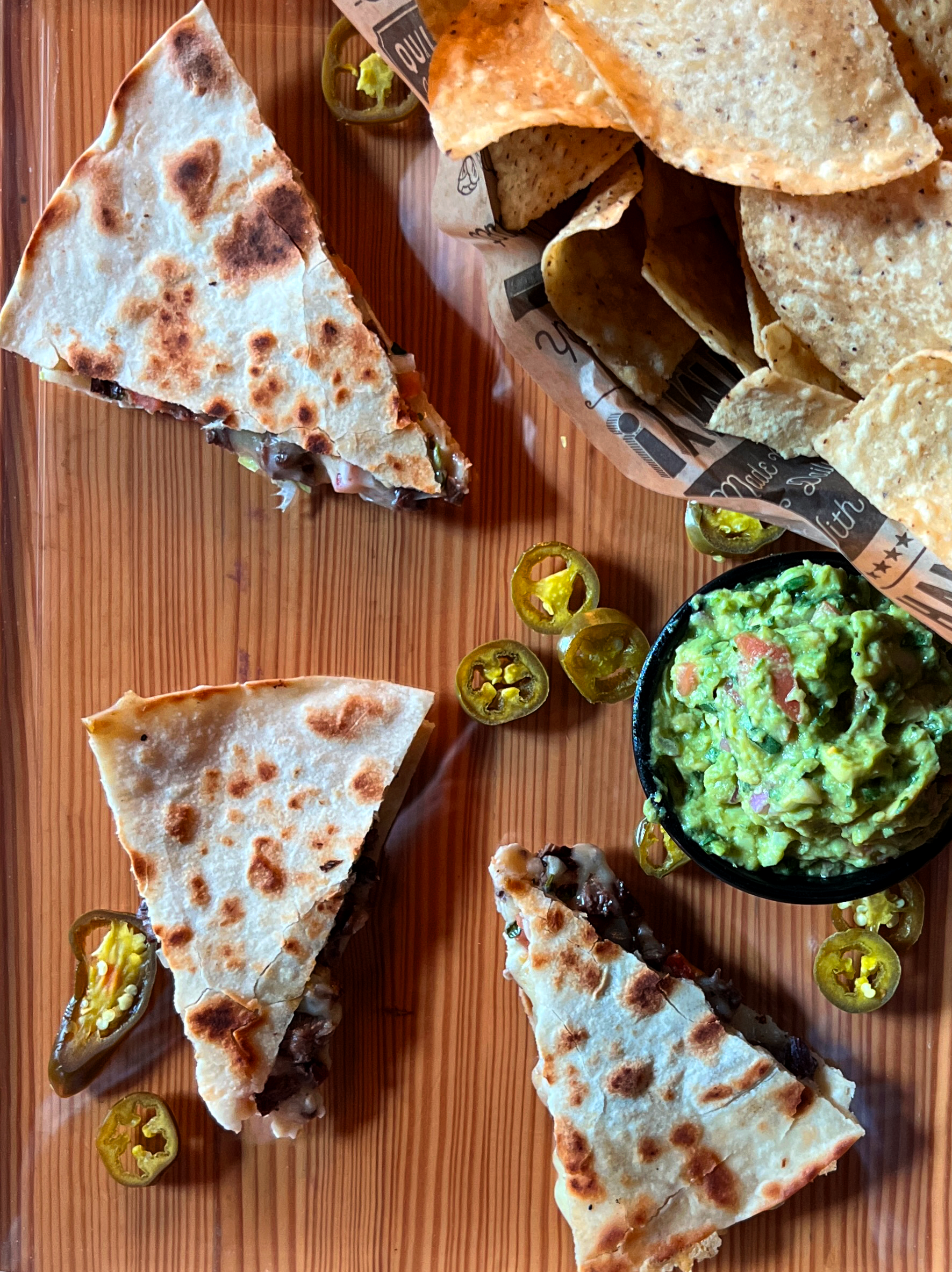 Quesadilla slices with guacamole, tortilla chips, and sliced jalapeños on a wooden table.