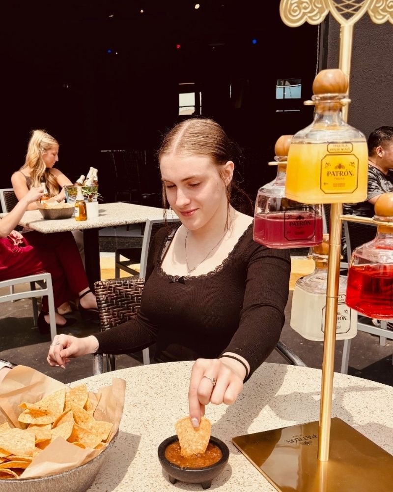 Young woman dipping a chip into red salsa at a restaurant table, with a basket of chips nearby and a display of bottles of sauce behind her.