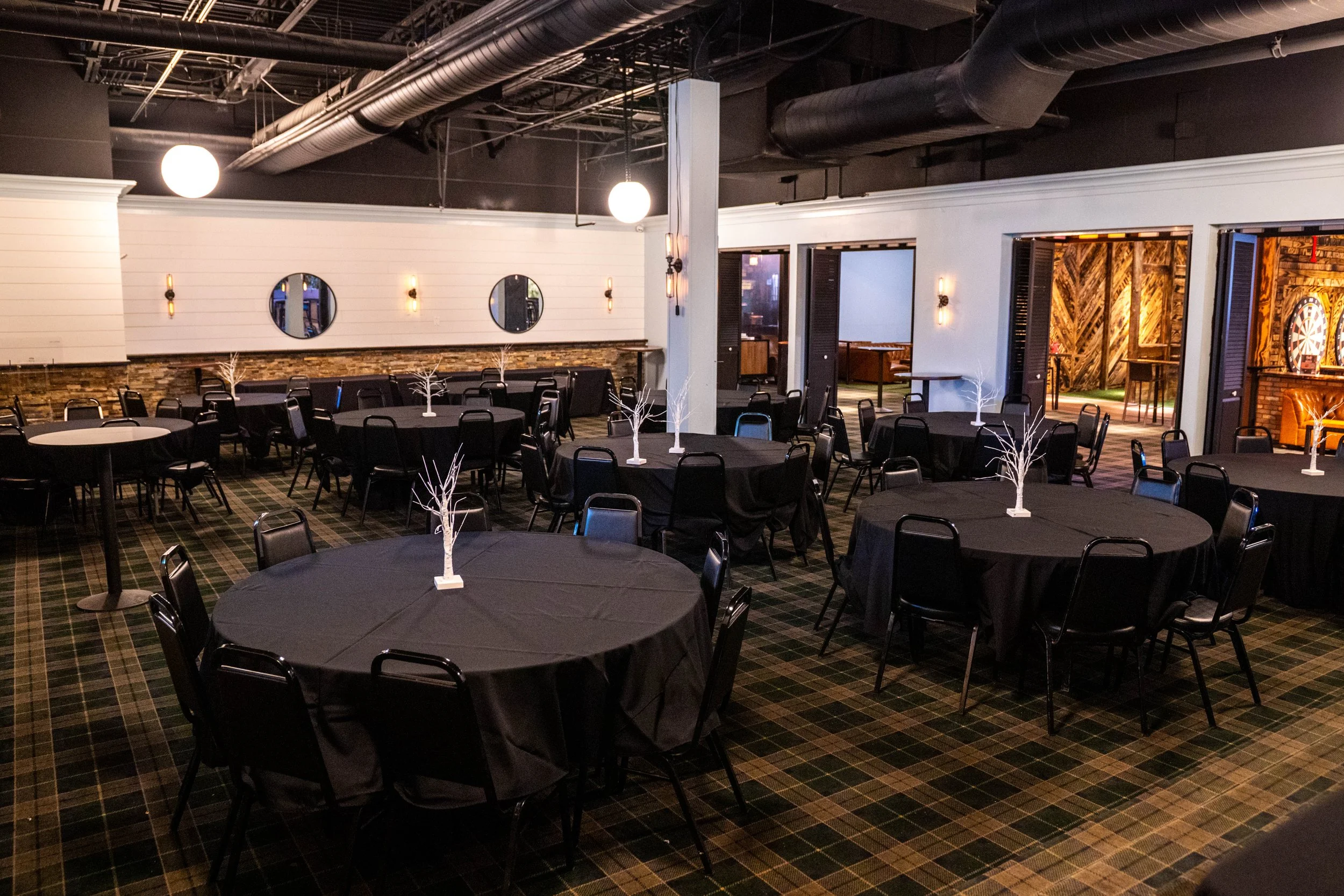 Interior of an event space with round tables and black chairs, featuring a checkered carpet and multiple wall mirrors. A screen displaying "HUB Stadium" is visible.