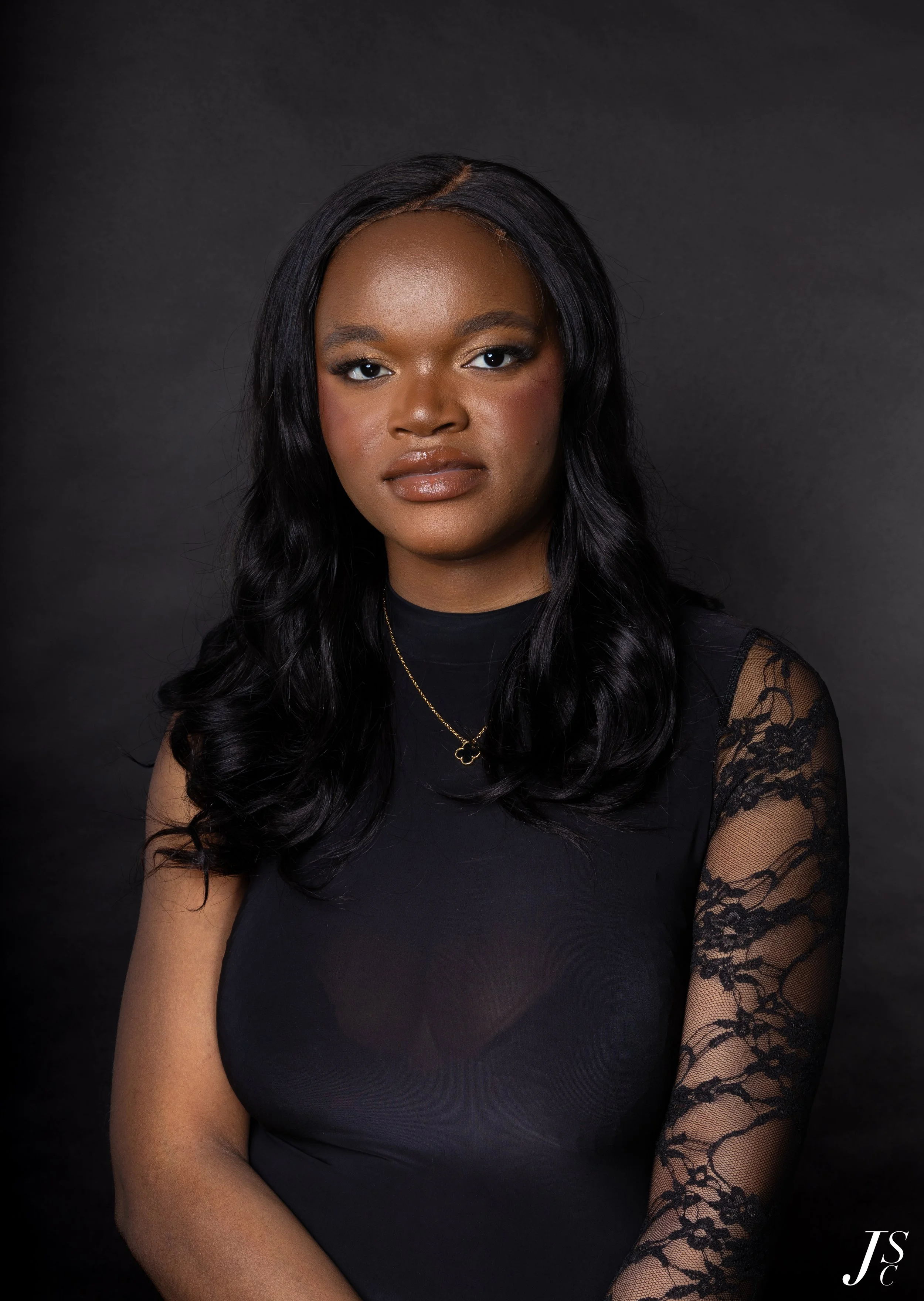 Portrait of a young African American woman with long, wavy black hair, wearing a black top with lace sleeves and a gold necklace, standing against a dark background.