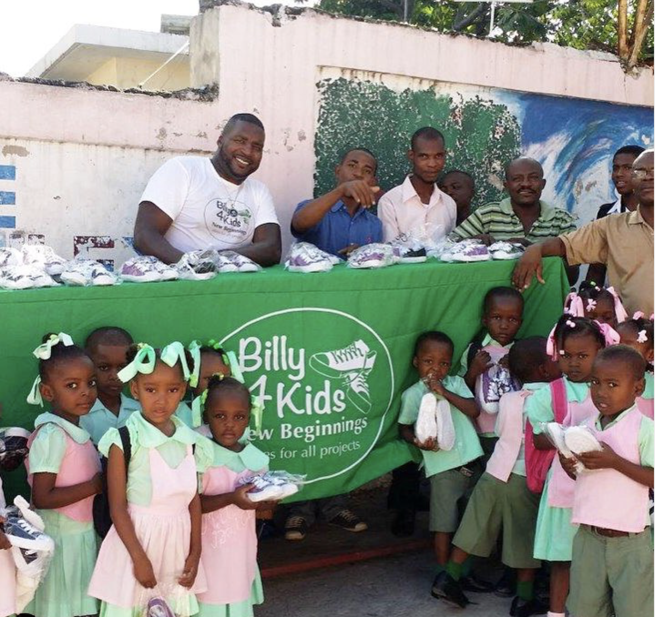 Children in school uniforms and smiling adults gathered around a table with a green "Billy 4 Kids, New Beginnings, Thanks for All Projects" banner, during an outdoor shoe donation event.
