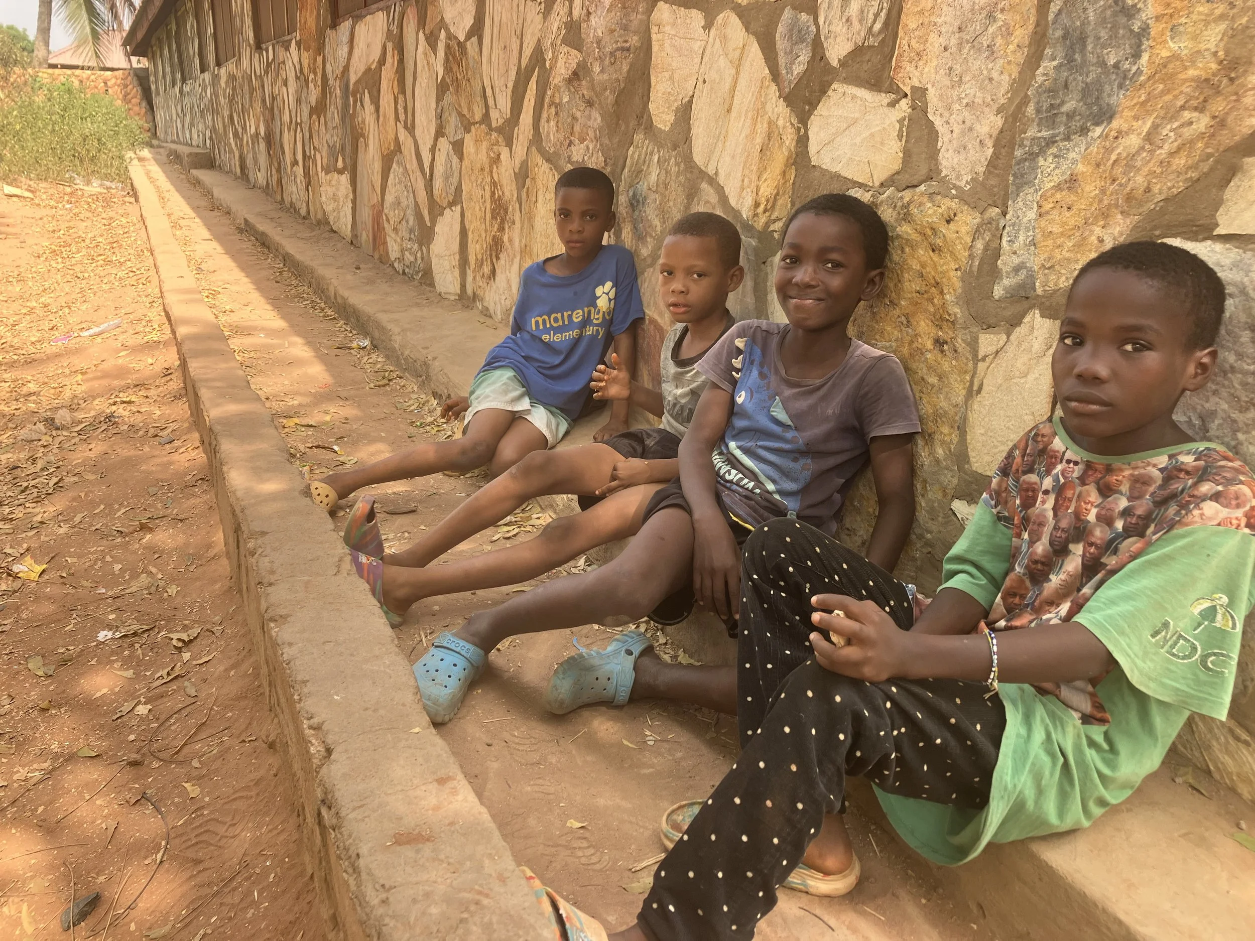 Four young boys sitting on a low concrete ledge in front of a stone wall on a dirt ground, wearing donated shoes from Billy4Kids; two are smiling while two have neutral expressions, highlighting the nonprofit’s global impact.