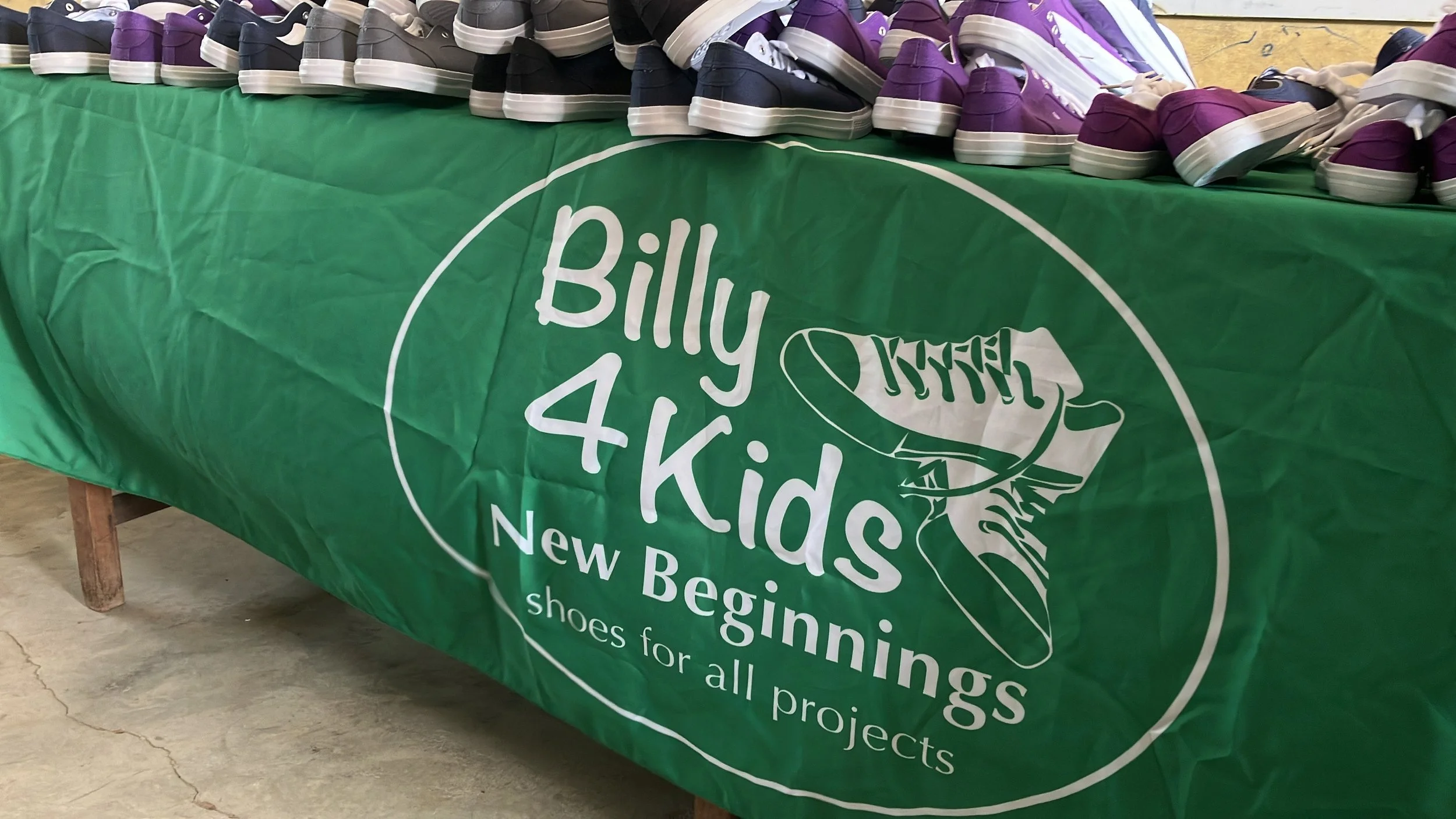 Table with a green banner reading "Billy 4 Kids, New Beginnings, shoes for all projects" and a display of purple, black, and gray sneakers on top.