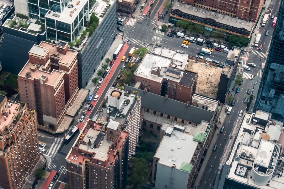 Aerial view of urban cityscape with streets, parked cars, and multiple buildings, including modern and older structures. Visible crosswalks and traffic on roads.