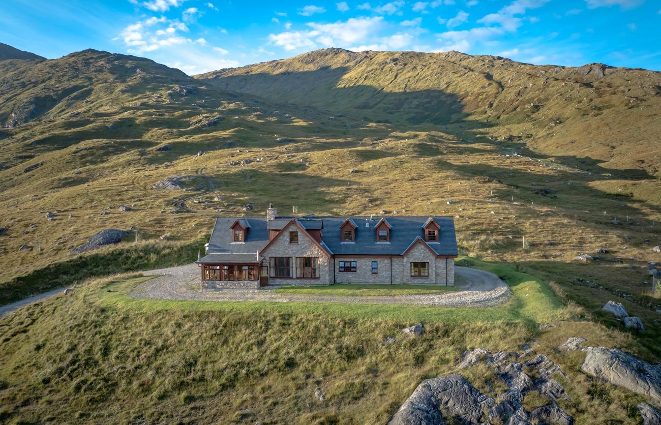 A large stone house with a dark gray roof and multiple windows is situated on a grassy hill in a mountainous landscape.