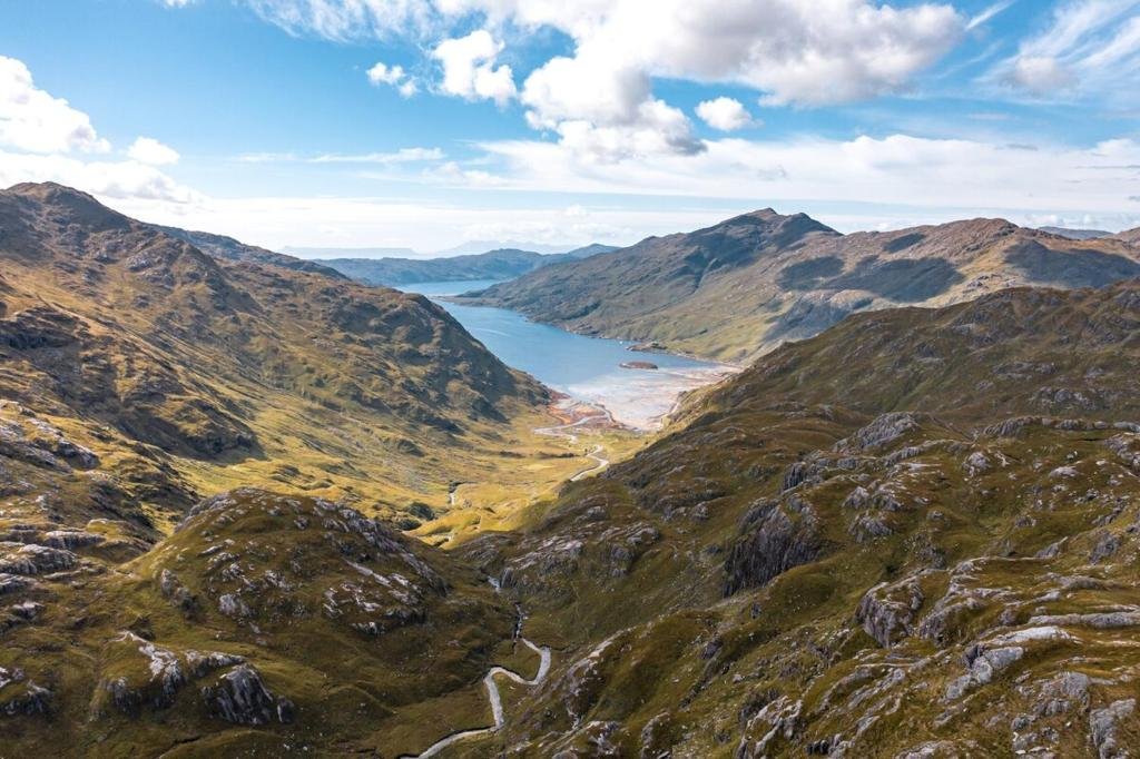 A scenic view of a valley with green hills and mountains, a winding river, and a large lake in the distance under a partly cloudy sky.
