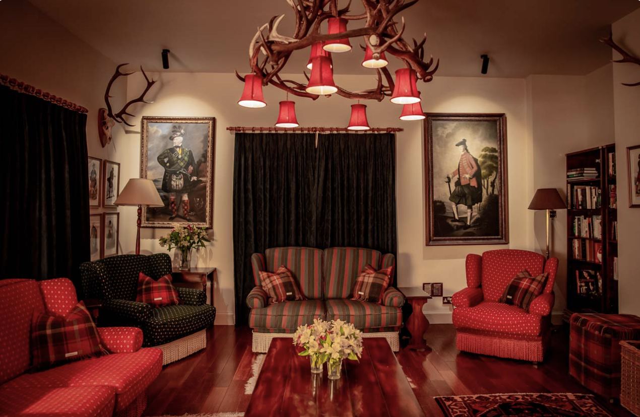 Living room decorated with red and black plaid furniture, framed portraits of historical figures, a floral centerpiece on a wooden coffee table, and antler chandelier with red lampshades.
