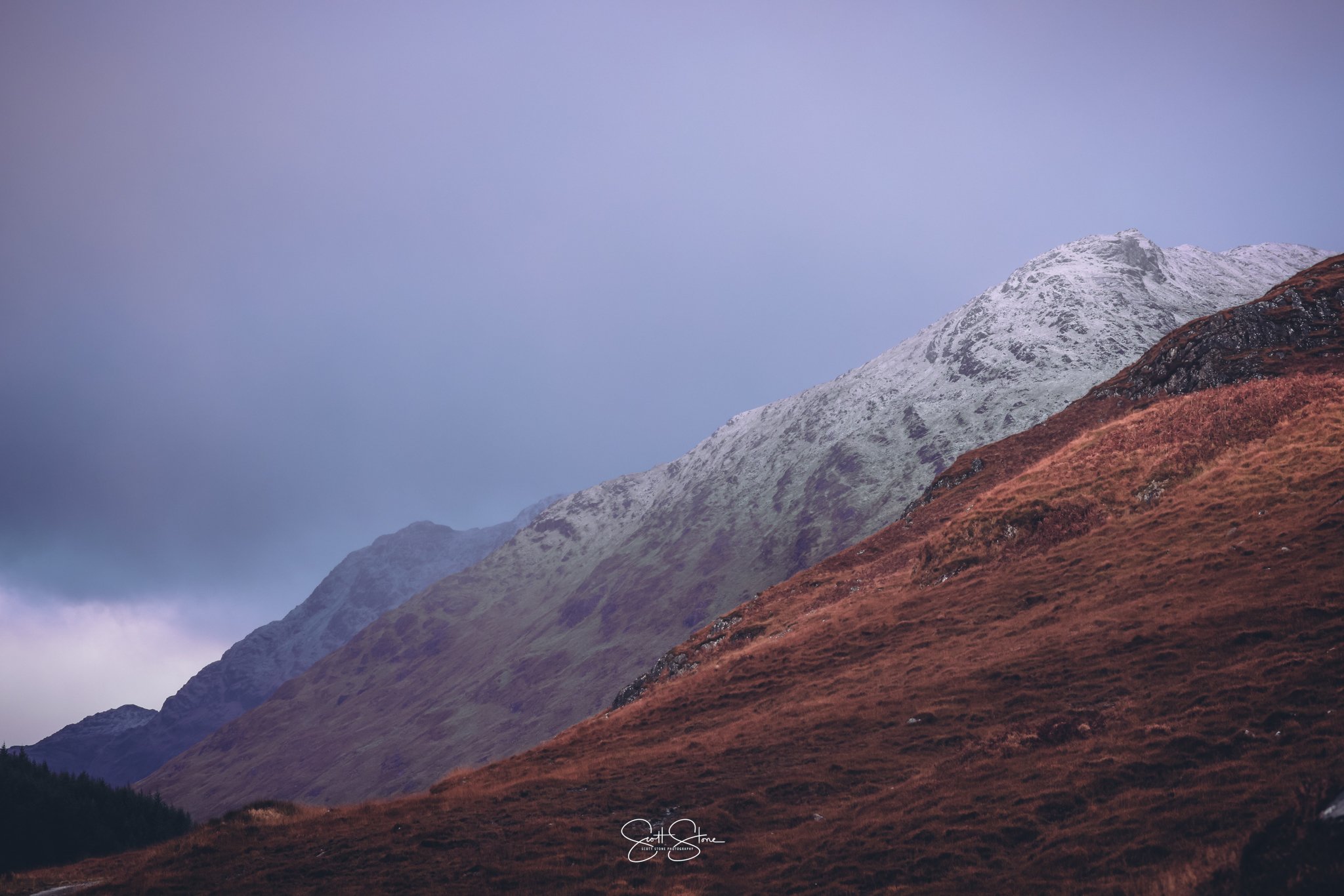 Snow-capped mountains next to reddish-brown hills under a cloudy sky.