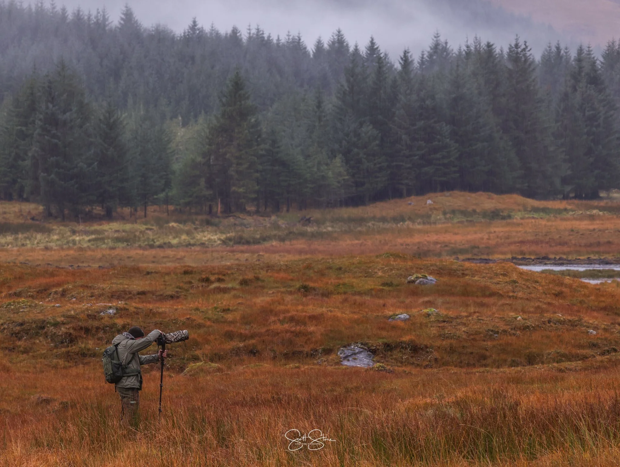 A person with a backpack and outdoor clothing, standing in a grassy field with a camera mounted on a tripod, taking photos of the landscape including distant trees and a cloudy sky.