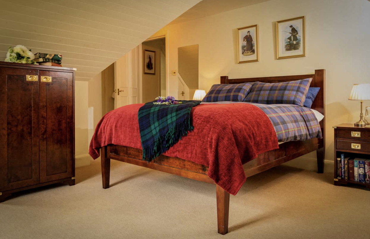 A cozy bedroom with a wooden bed frame, plaid bedding, red coverlet, and a dark green tartan blanket. There are two nightstands with lamps, framed artwork above the bed, and a wardrobe with books and flowers on top.