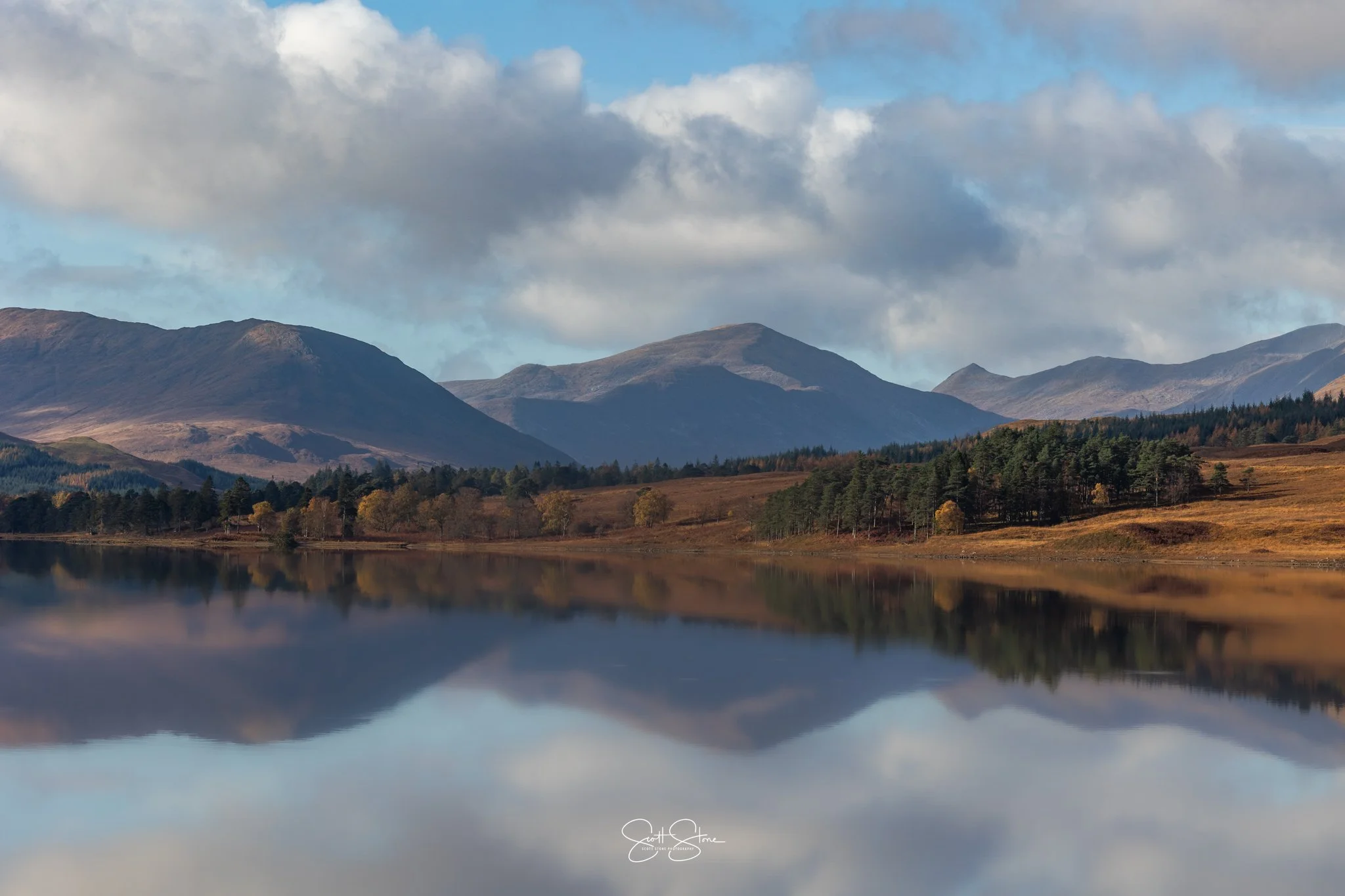 A serene landscape featuring a mountain range in the background, a calm lake reflecting the mountains and sky, and a forested area with trees along the shoreline.