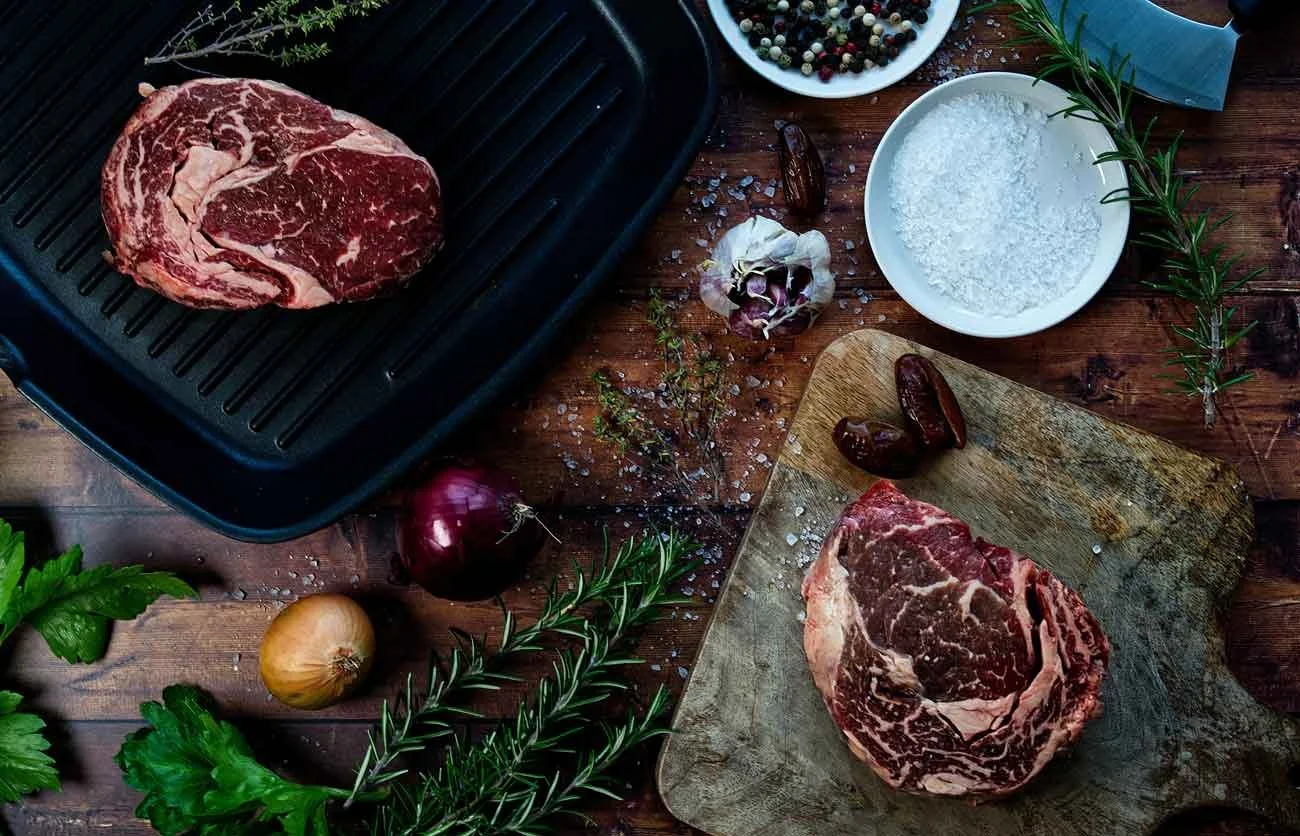 Raw beef steaks on a wooden cutting board and black grill pan, surrounded by herbs, onions, garlic, and bowls of salt and peppercorns.