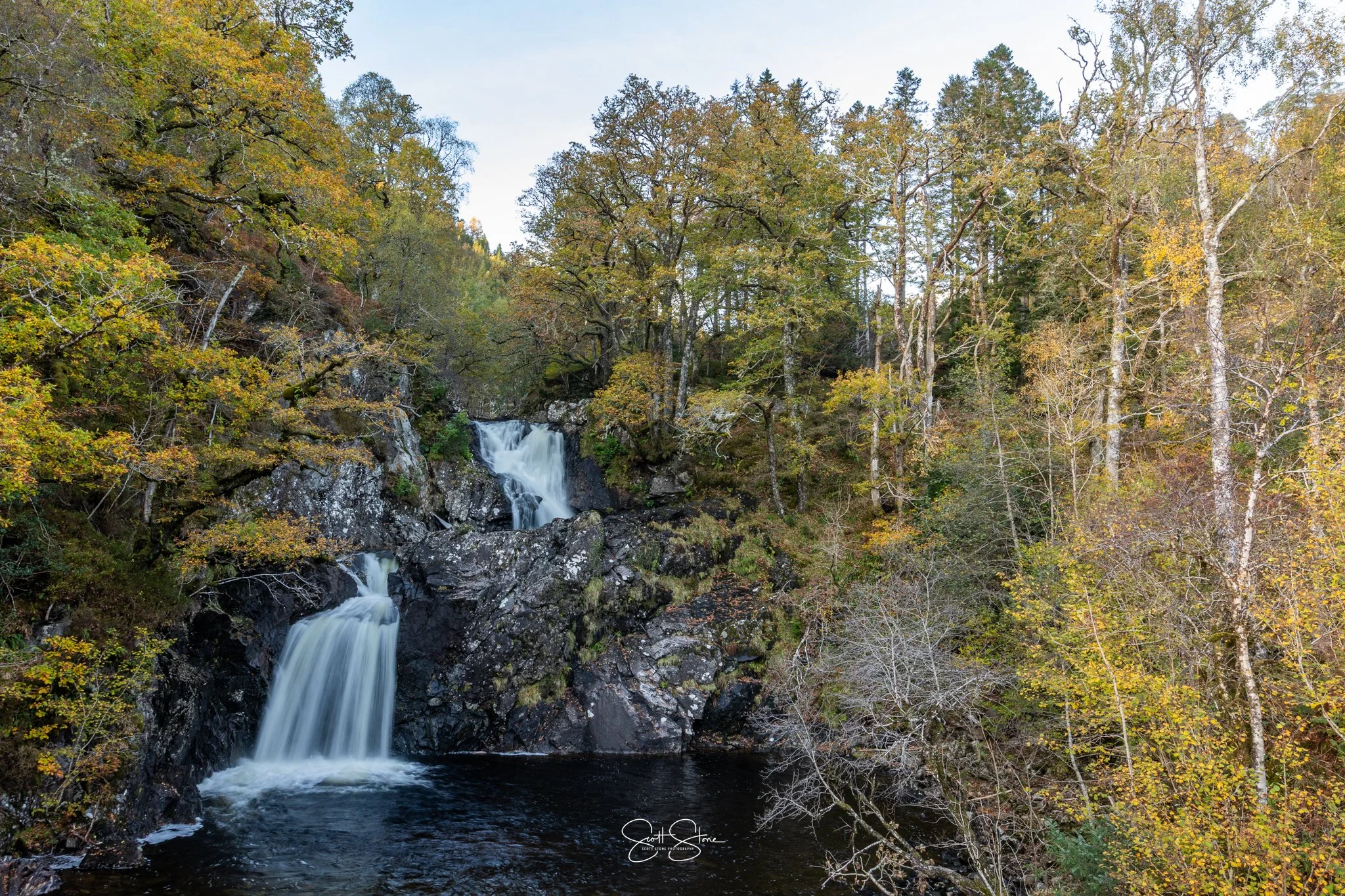 A waterfall flowing over rocks in a forest with trees showing autumn foliage.