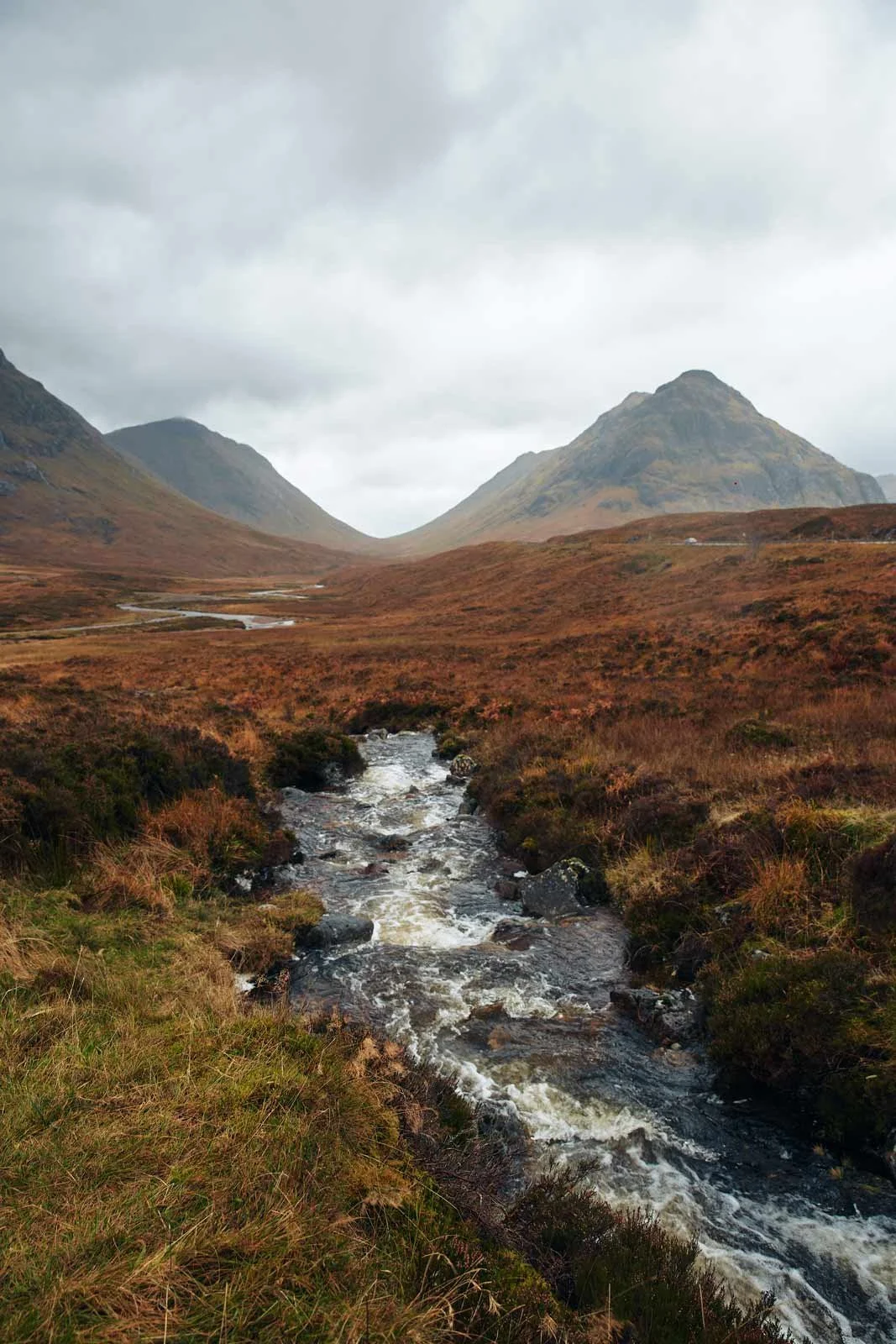 A mountain landscape with a cloudy sky, a small stream flowing through grassy and shrub-covered terrain, and distant mountains in the background.
