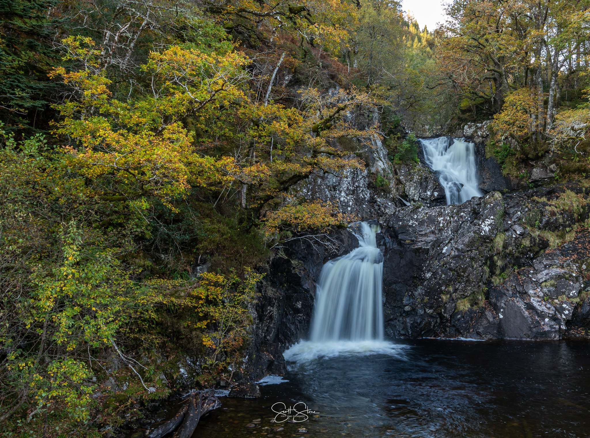 A two-tier waterfall flowing over rocks surrounded by autumn foliage in a forest