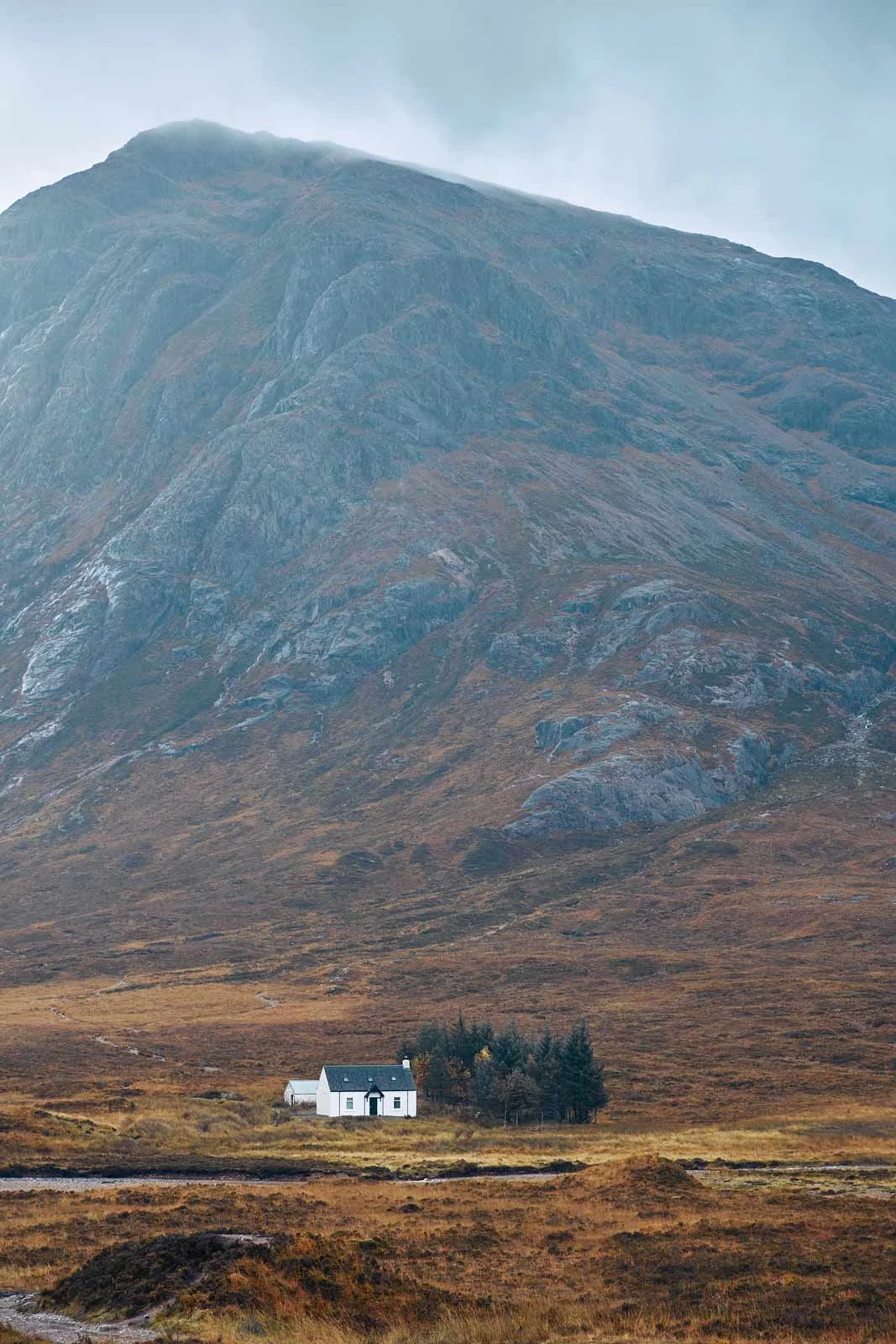 A small white house with a dark roof and chimney situated in a grassy, brown landscape near a cluster of trees. In the background, a large, rugged mountain with a partly cloudy sky above.