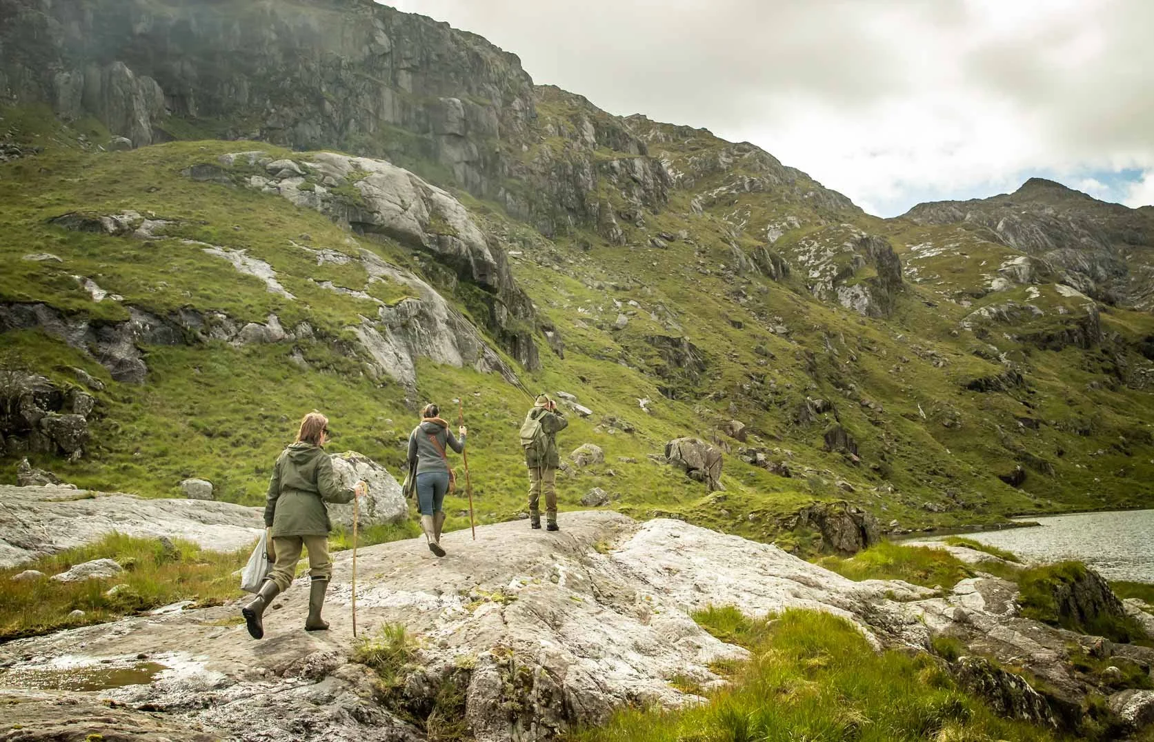 Three people hiking along a rocky trail near a lake with green mountains in the background.