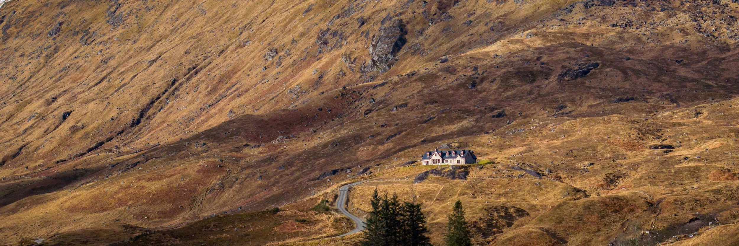 A house situated on a hillside in a mountainous landscape with dry grass, rocks, and a winding road leading up to it, under a clear sky.