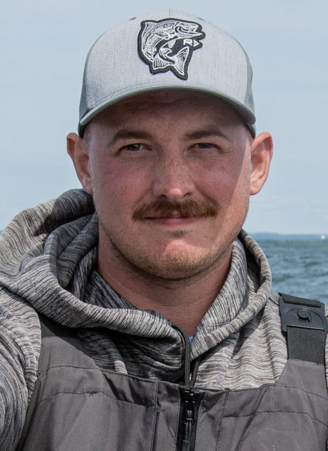 A man with short hair, wearing a gray baseball cap with a fish logo, a gray hoodie, and a gray vest, standing outdoors near a body of water.