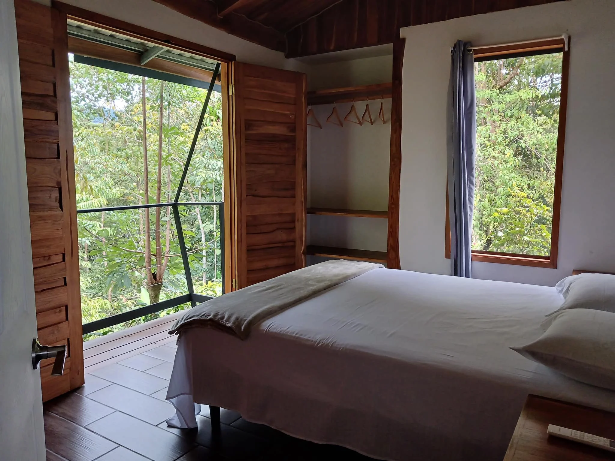 A cozy bedroom with a large bed covered in white sheets and pillows, wooden window frames, and an open balcony door revealing lush green trees outside.