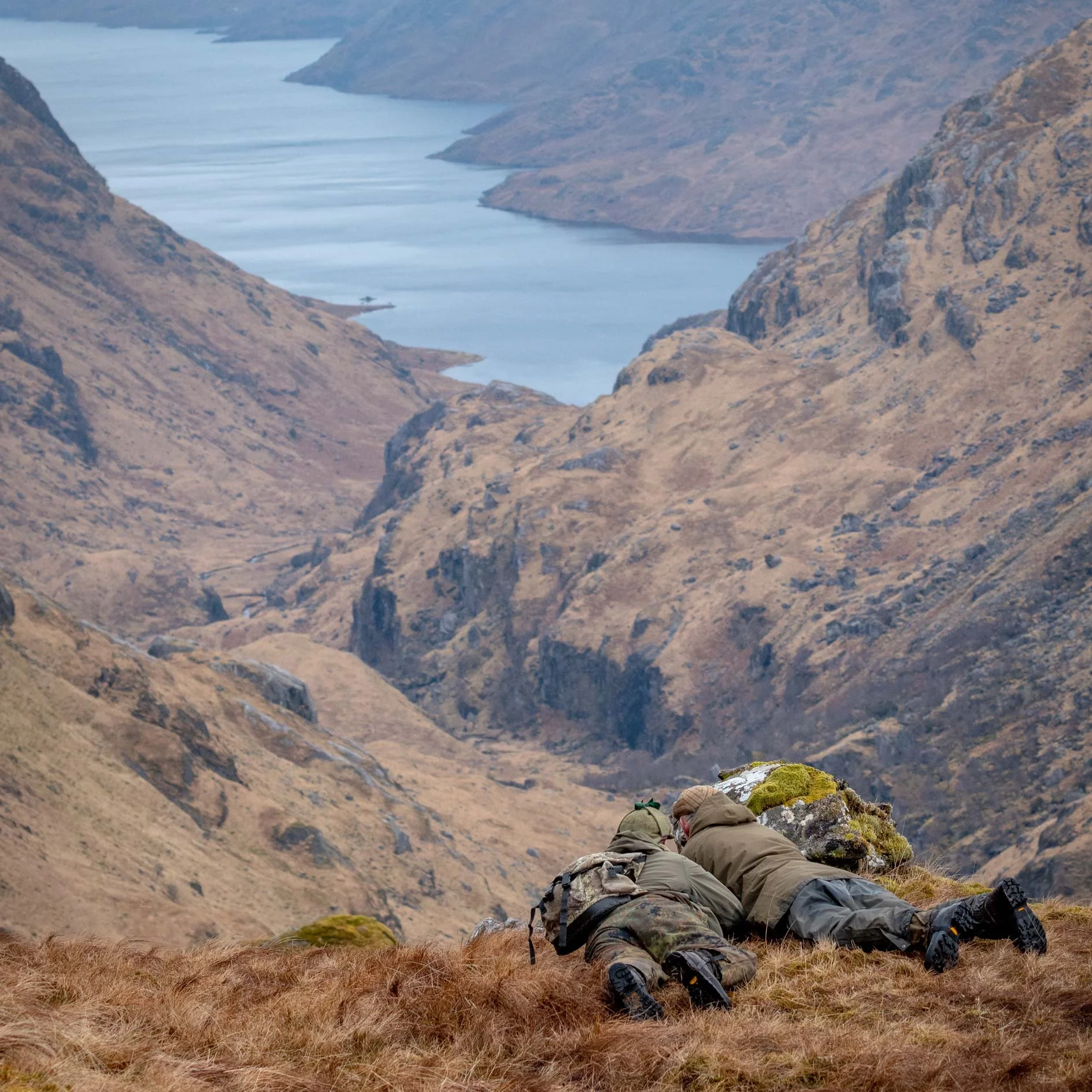Two hikers in outdoor gear lying on their stomachs on grassy terrain, looking out over a scenic landscape of mountain valleys, a river, and a lake in the distance.