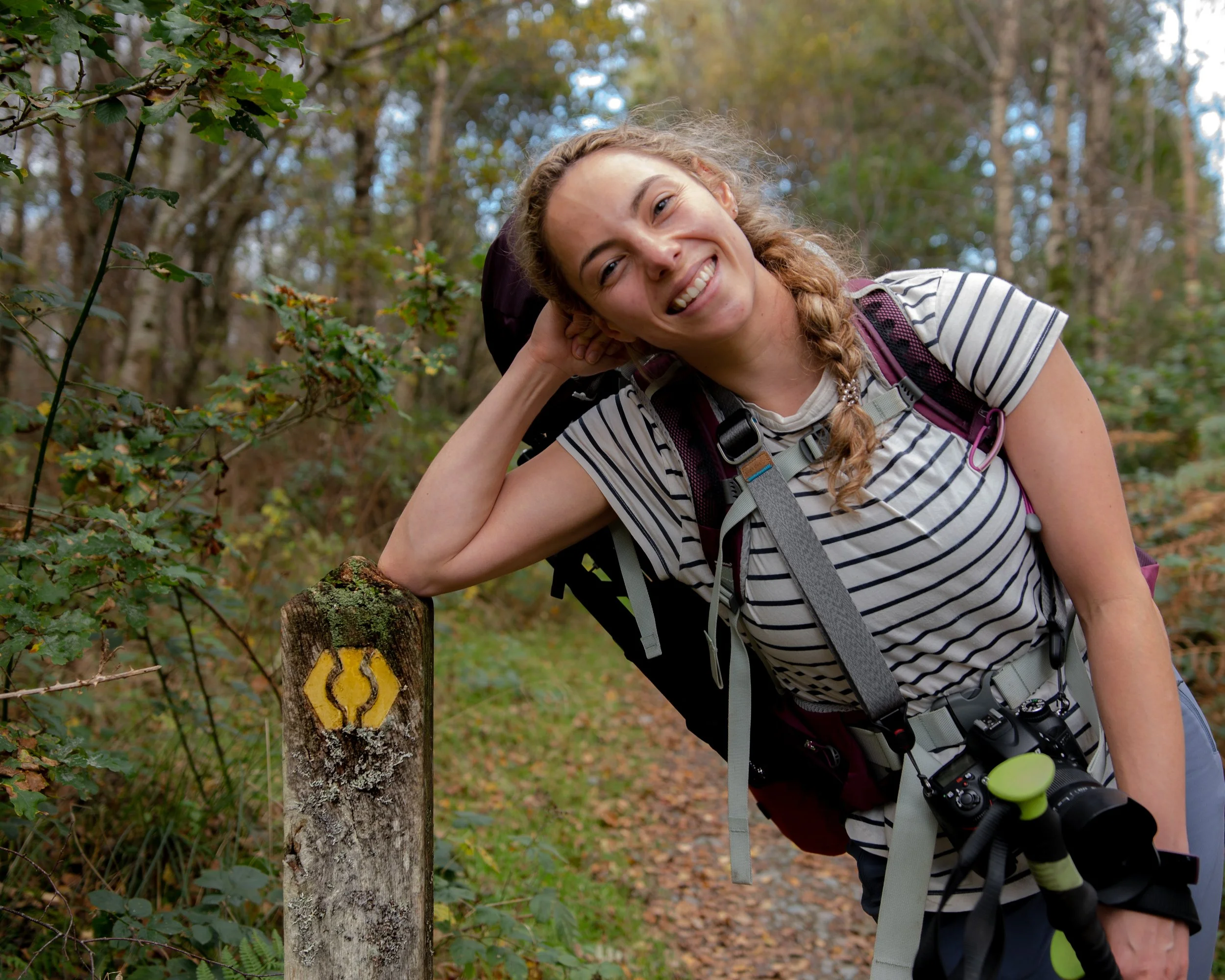 A young woman smiling in a striped t-shirt outdoors in a wooded area, leaning on a trail marker with a yellow blaze, carrying a backpack and camera.