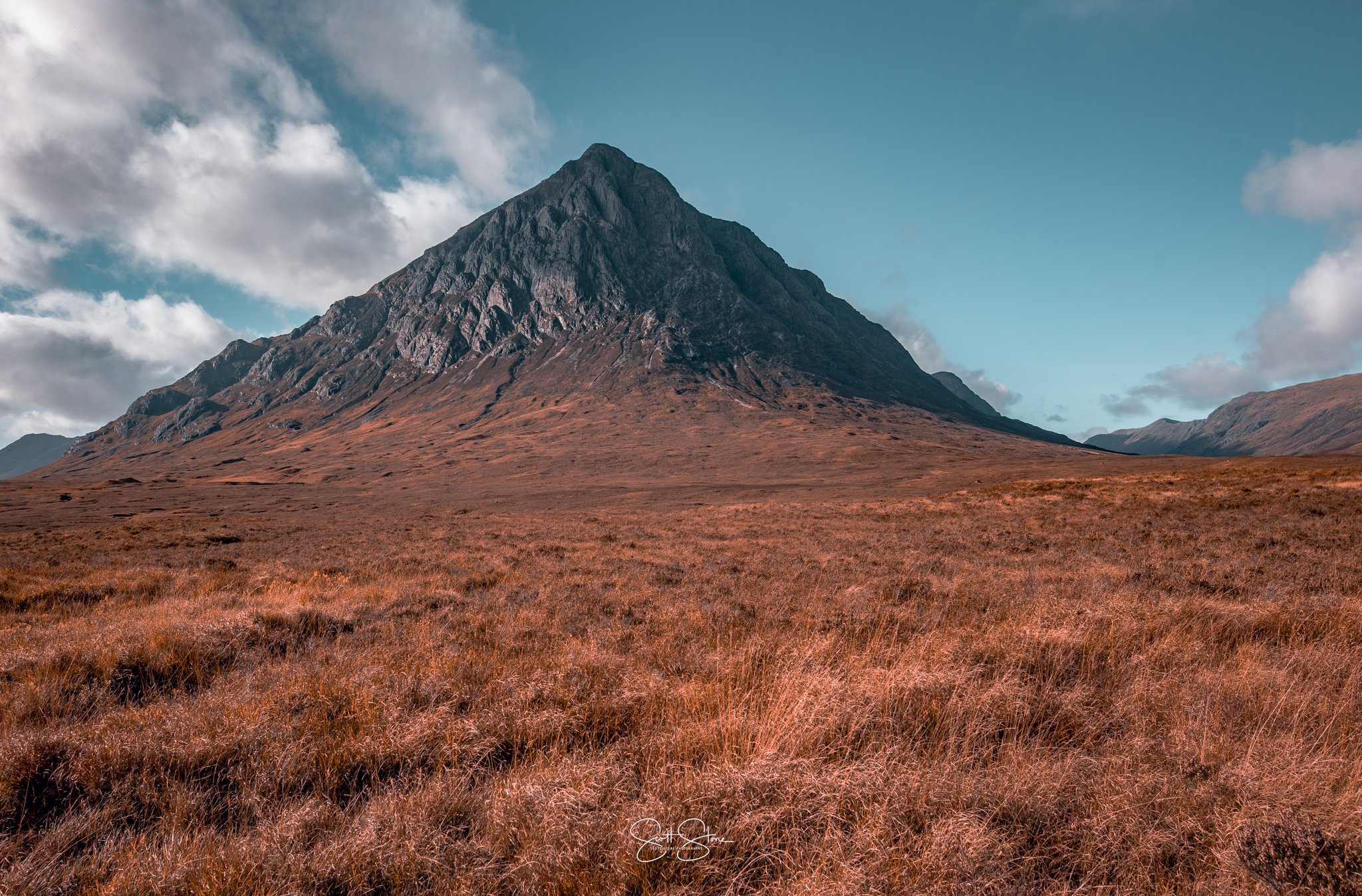 A large mountain peak rising over a vast open landscape with reddish-brown grass and a partly cloudy sky.