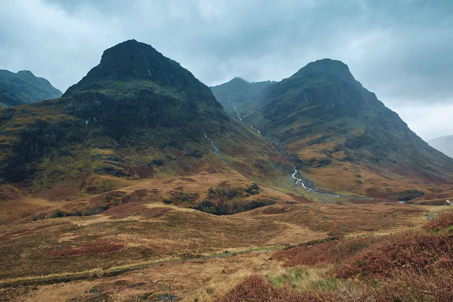 Scenic view of a rugged mountain landscape with two prominent peaks, a green valley, and a waterfall flowing down the mountainside under a cloudy sky.