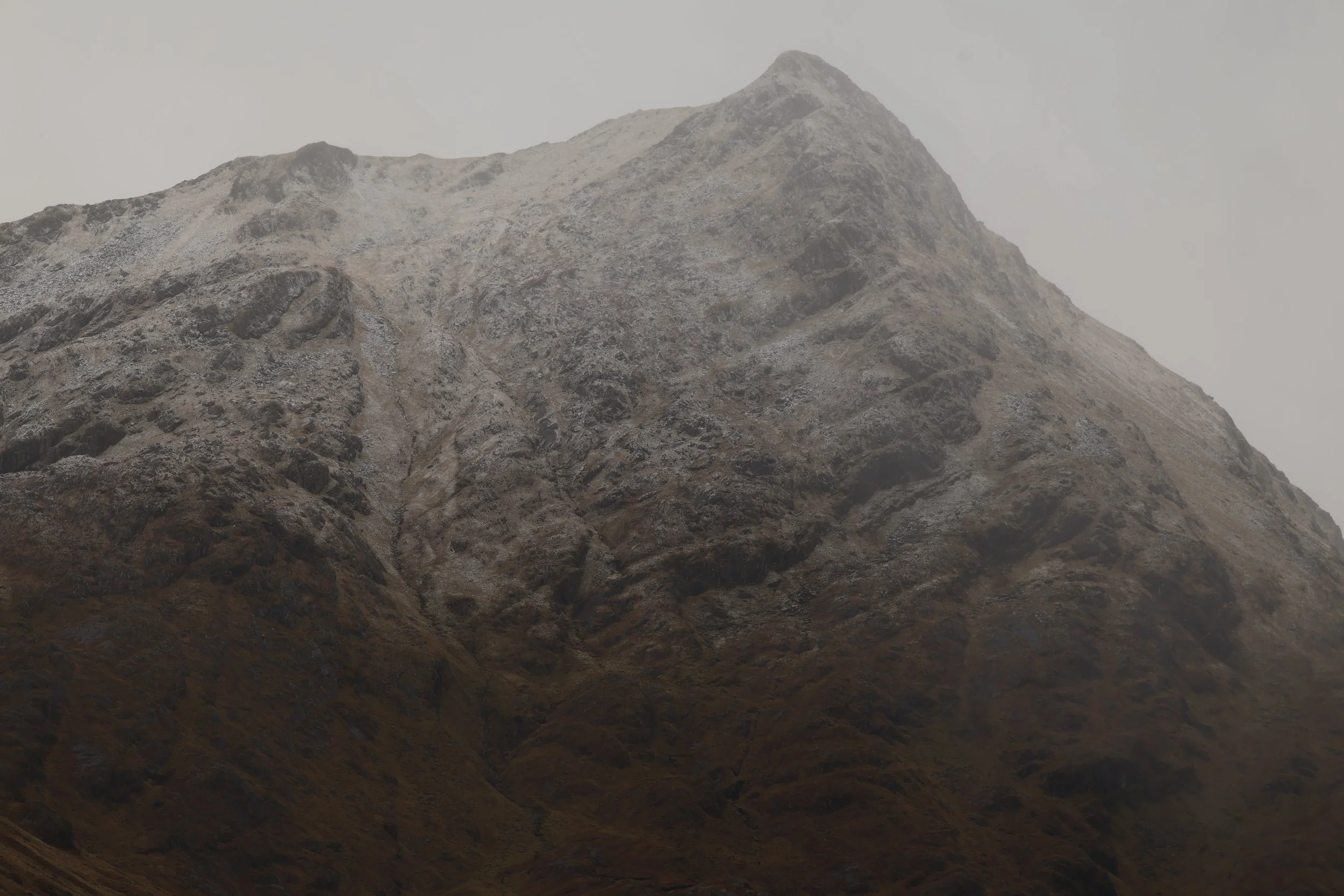 A rugged mountain with a rocky, uneven surface, partially covered in snow or frost, with a cloudy, overcast sky in the background.