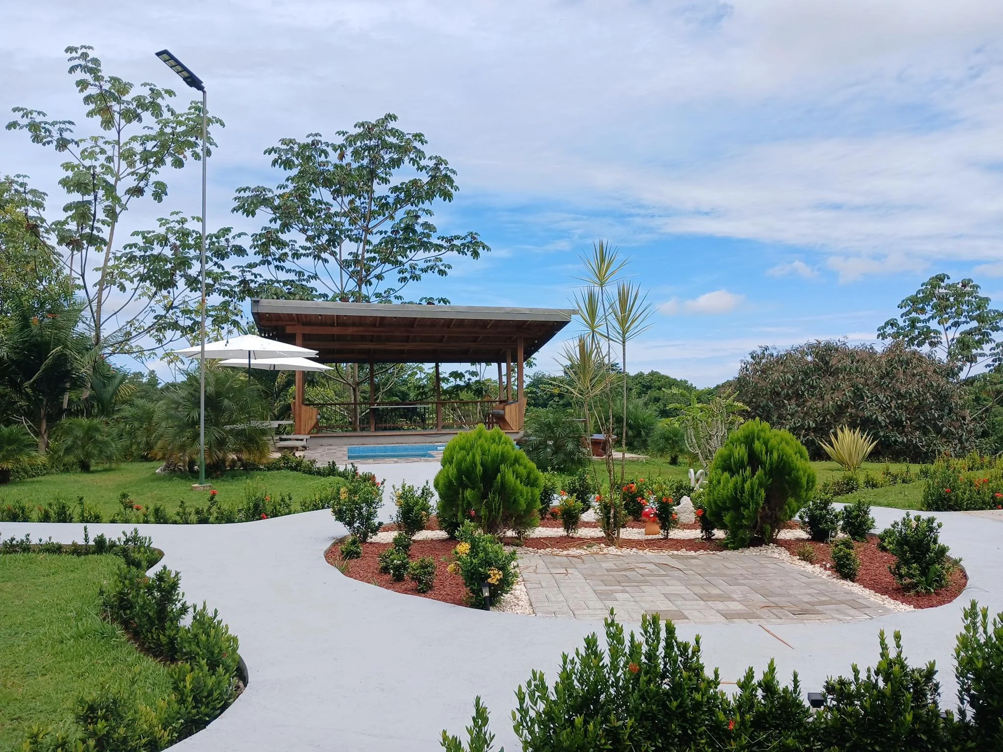 A landscaped backyard with a circular white pathway, lush green plants, small trees, a gazebo with seating, a swimming pool, and a blue sky with clouds.