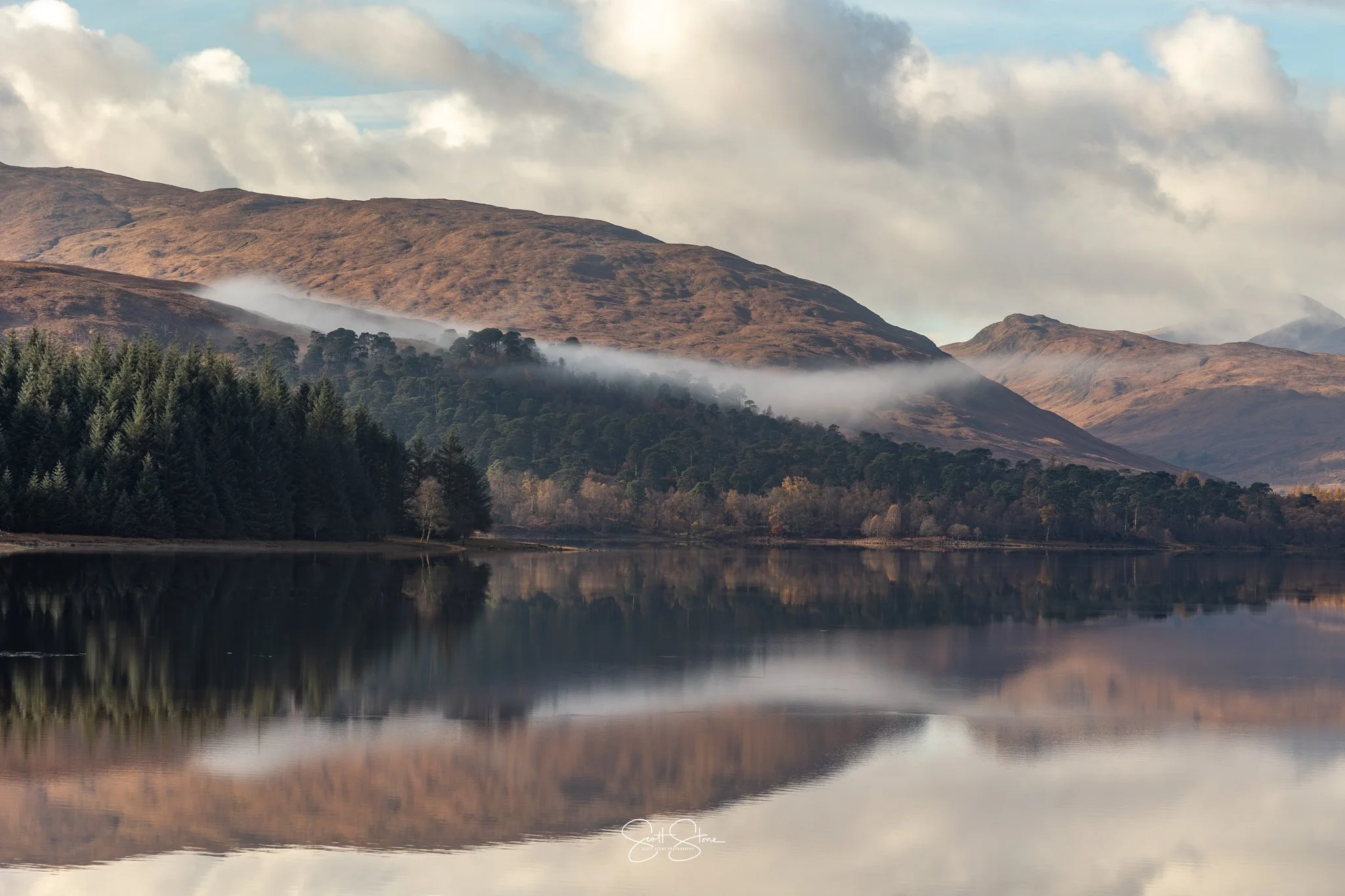 A calm lake reflecting surrounding mountains with brown and green vegetation, some clouds overhead, and a strip of fog or mist along the mountain slopes.