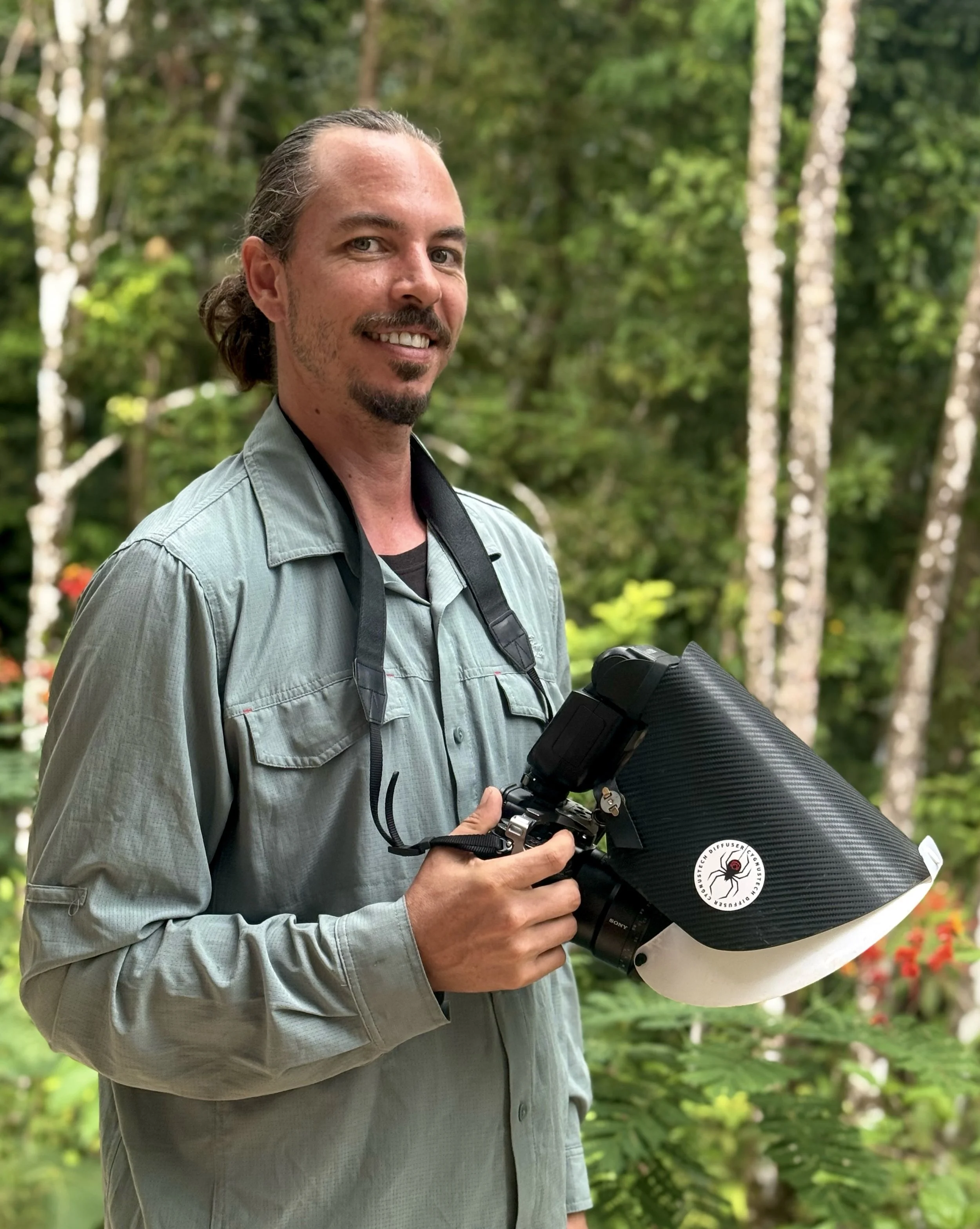 A man standing outdoors in a wooded area holding a camera with a large black attachment, wearing a light gray shirt and smiling.