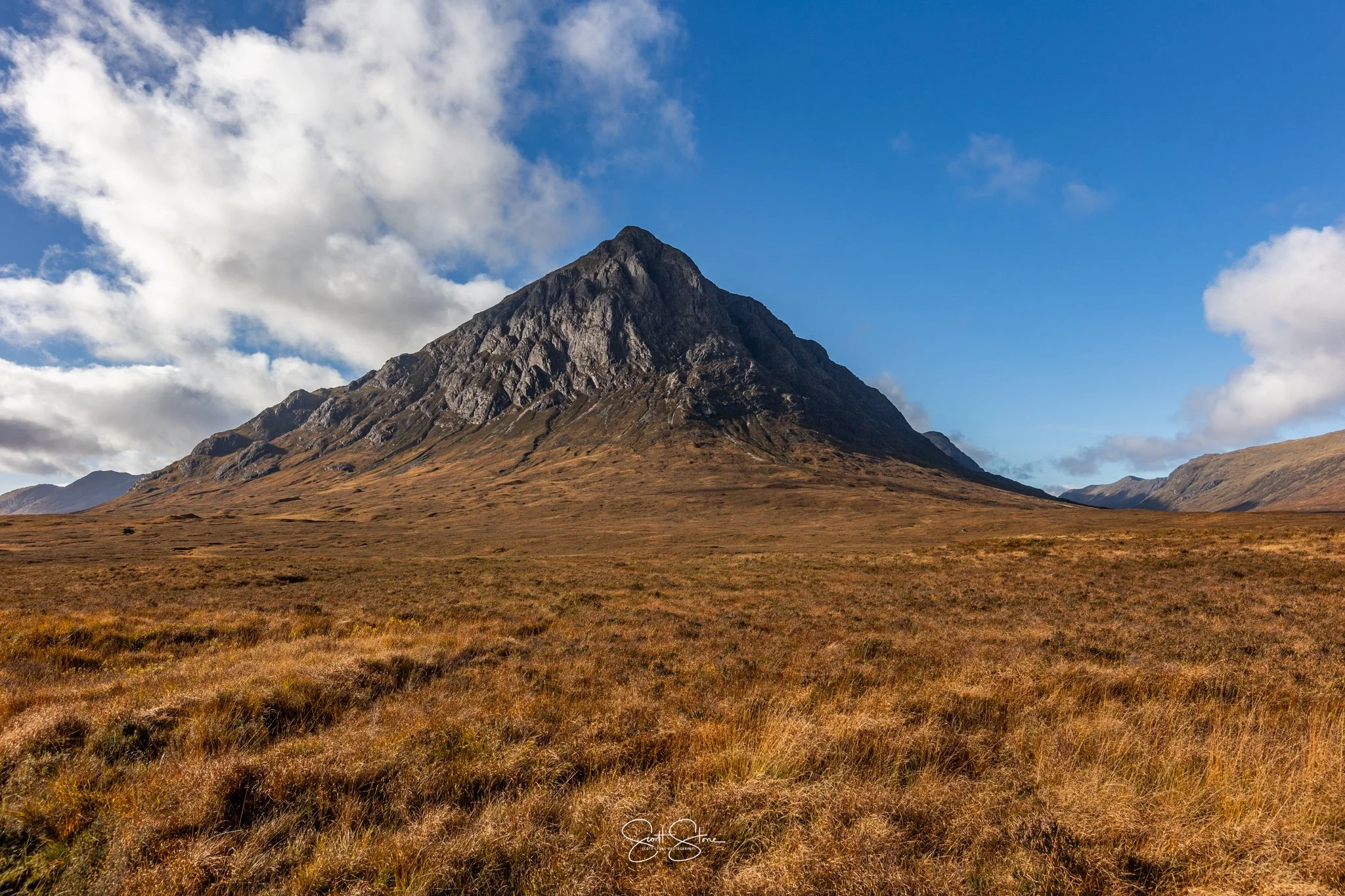A large, rugged mountain with steep slopes and rocky terrain, under a partly cloudy sky, in a vast open plain with golden-brown grass.