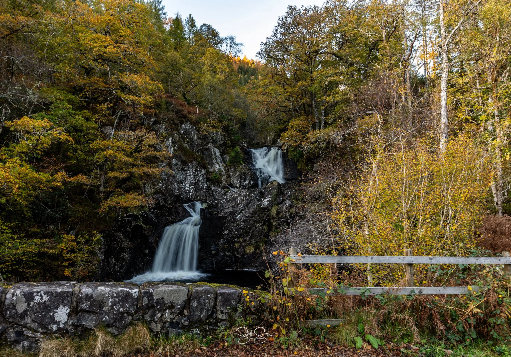 A waterfall cascading over rocks surrounded by trees with autumn foliage, a wooden fence in the foreground.