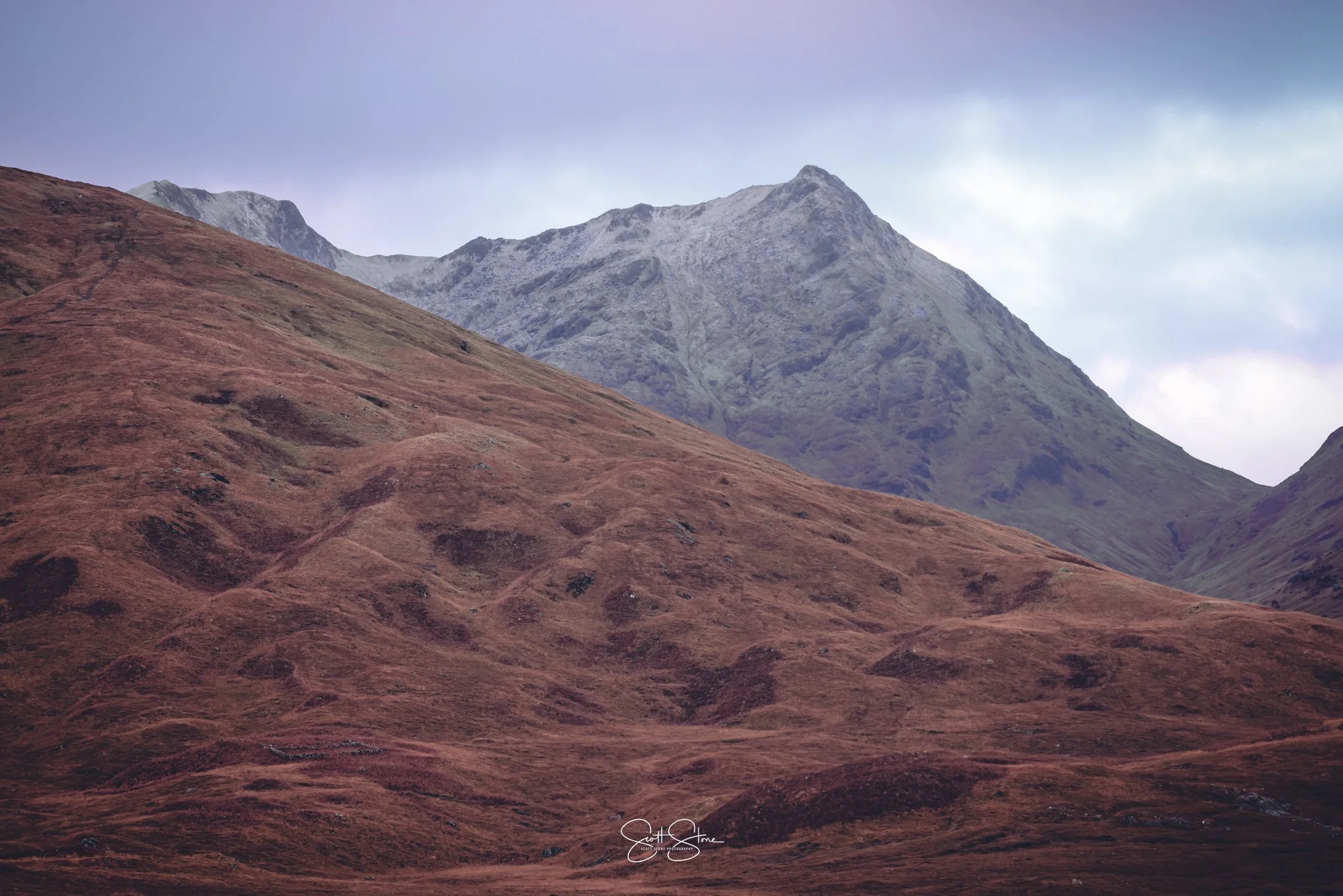 View of mountains with reddish-brown slopes in the foreground and rocky peaks in the background under a cloudy sky.