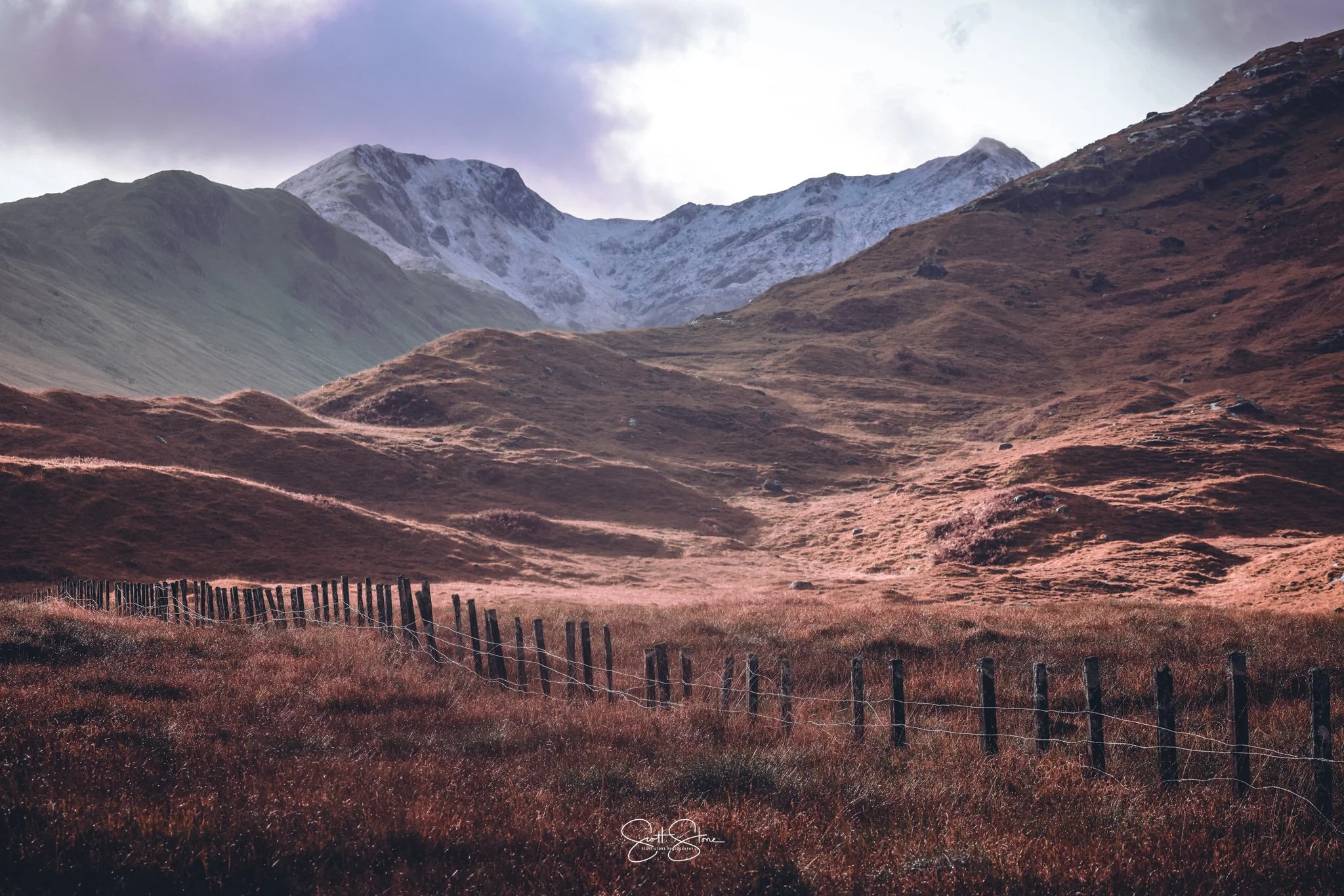 A scenic landscape of rolling hills with brown grass, a fenced path, and mountains with snow on their peaks in the background, under a cloudy sky.