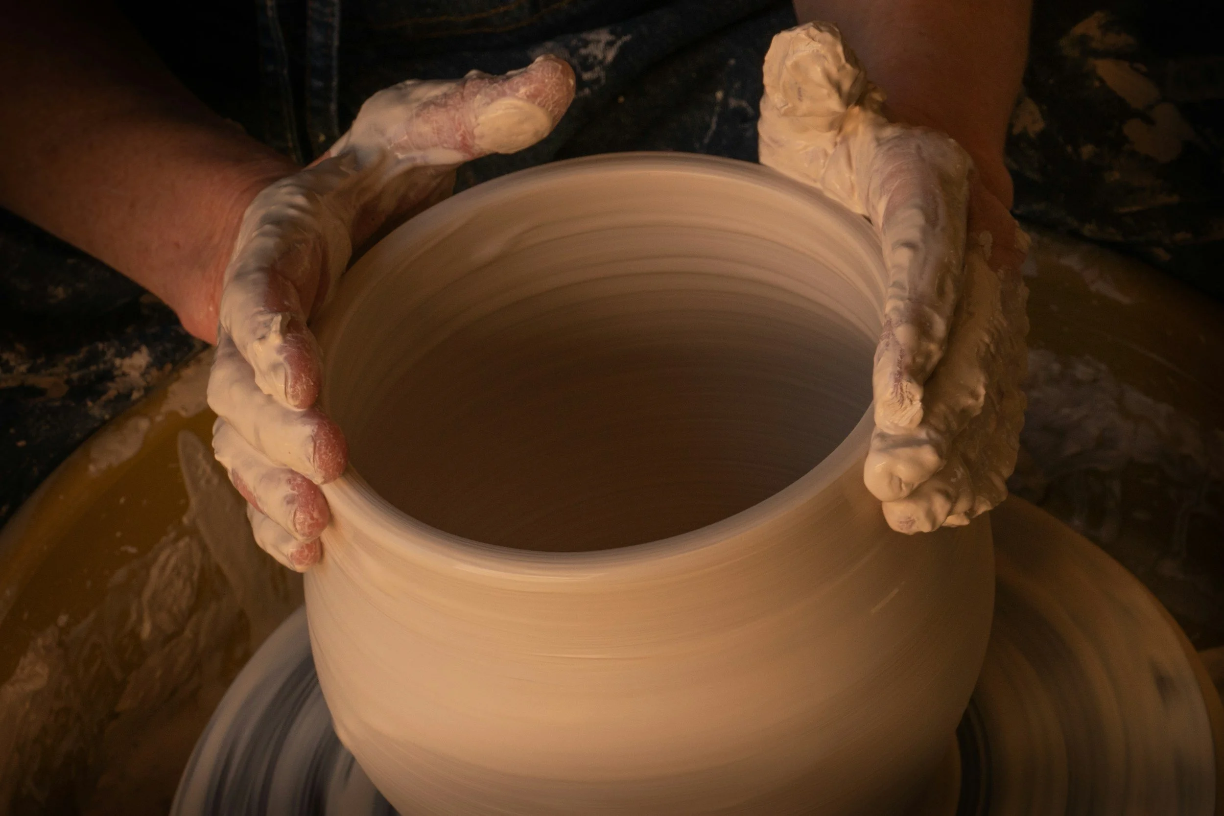 Hands shaping a large clay vase on a pottery wheel with wet clay on fingers and hands.