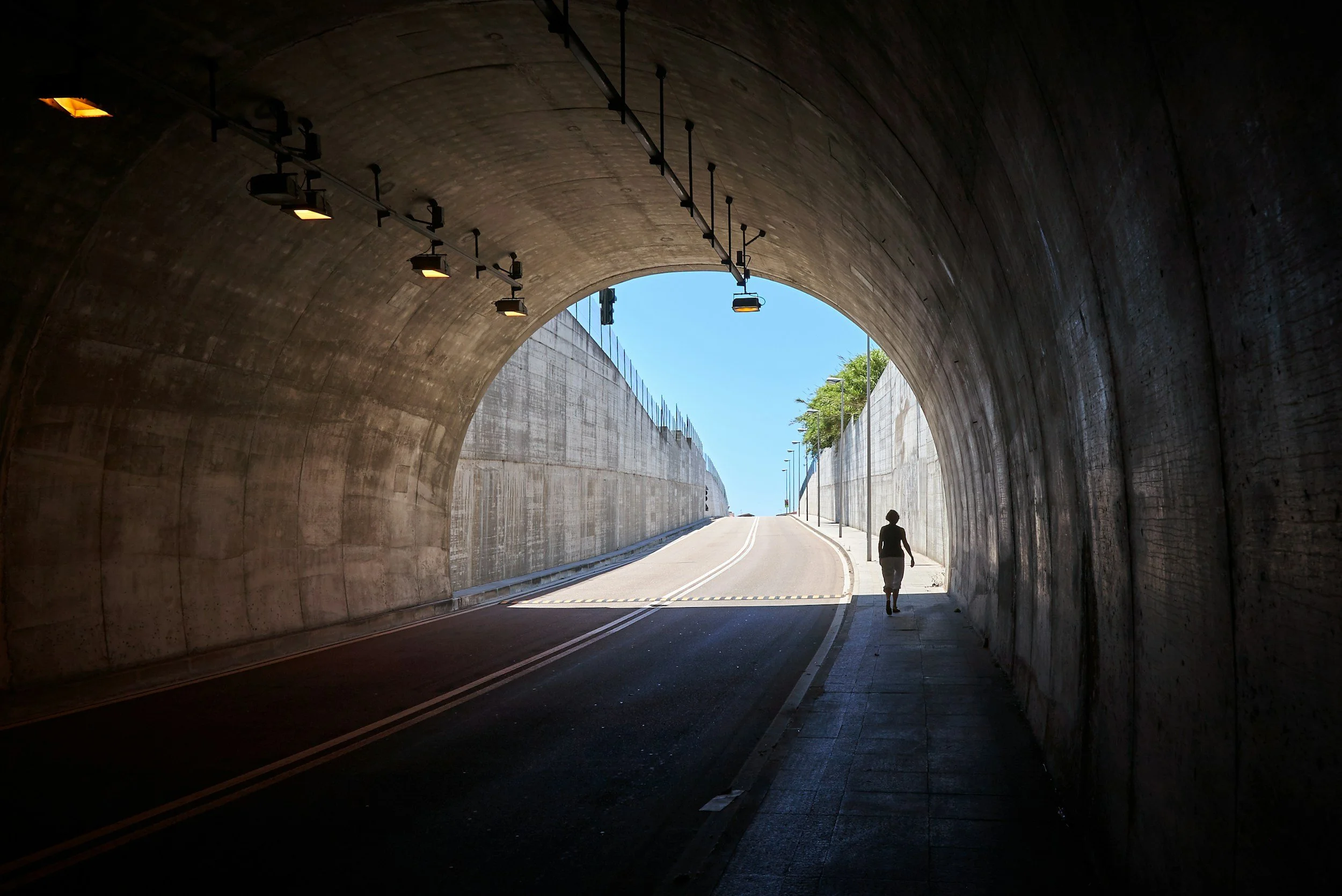 A person walks through a dark tunnel towards the bright open road outside, with clear blue sky and trees visible in the distance.