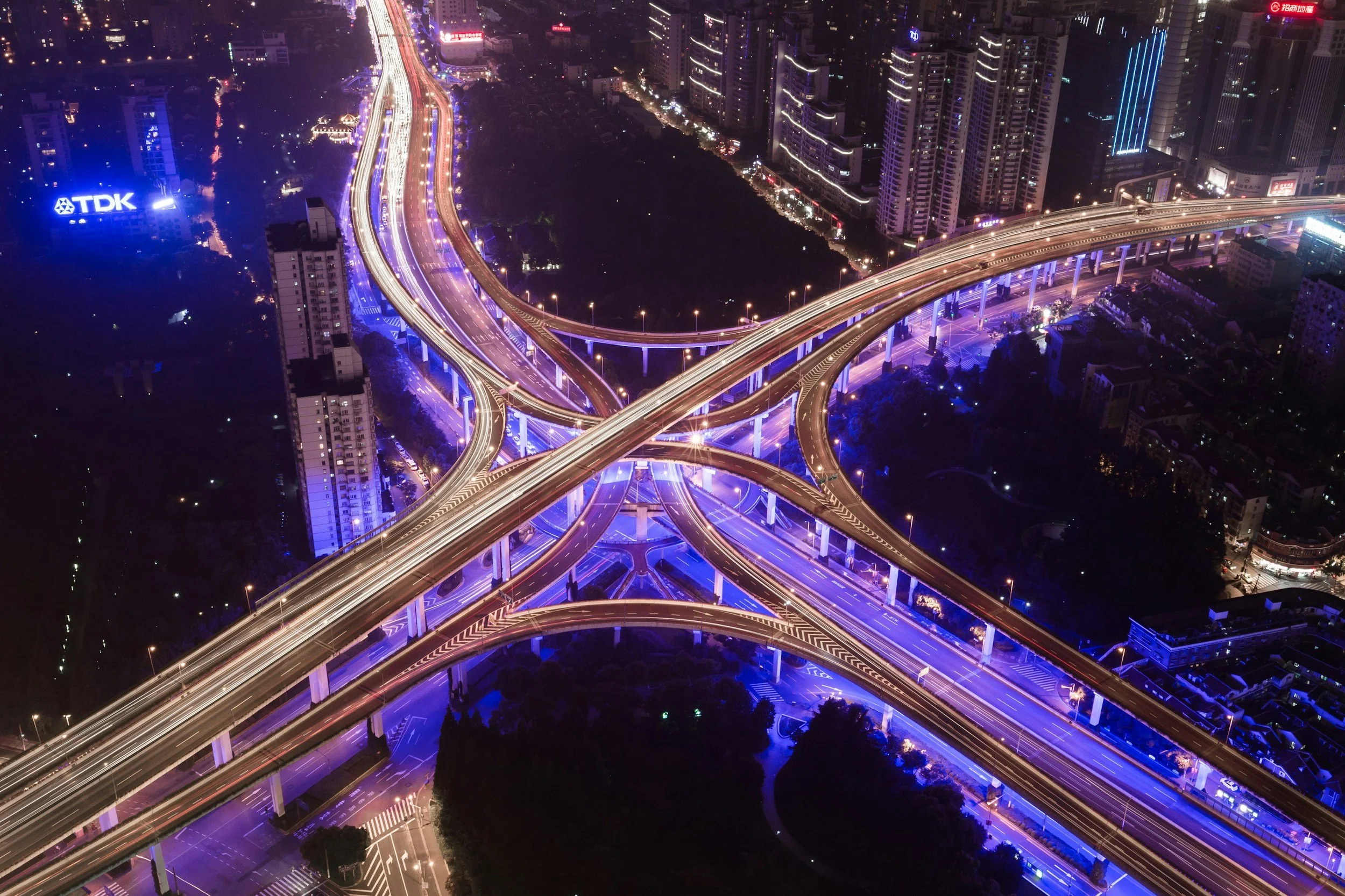 Aerial night view of a complex highway interchange with overpasses and bridges illuminated by multicolored city lights, surrounded by high-rise buildings and city streets.