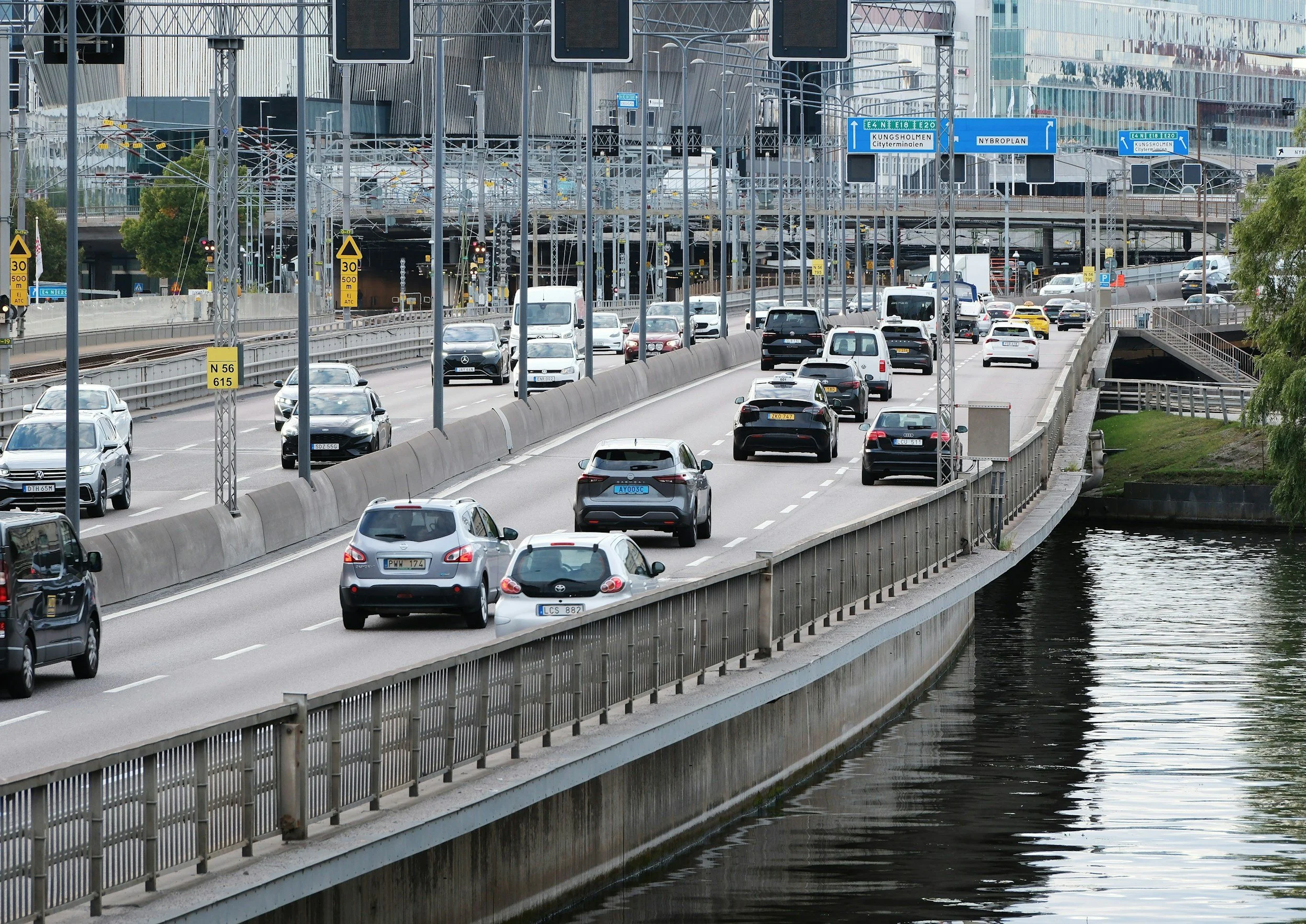 A busy multi-lane highway with cars and trucks driving beside a body of water under an overpass. Street signs and traffic signals are visible.