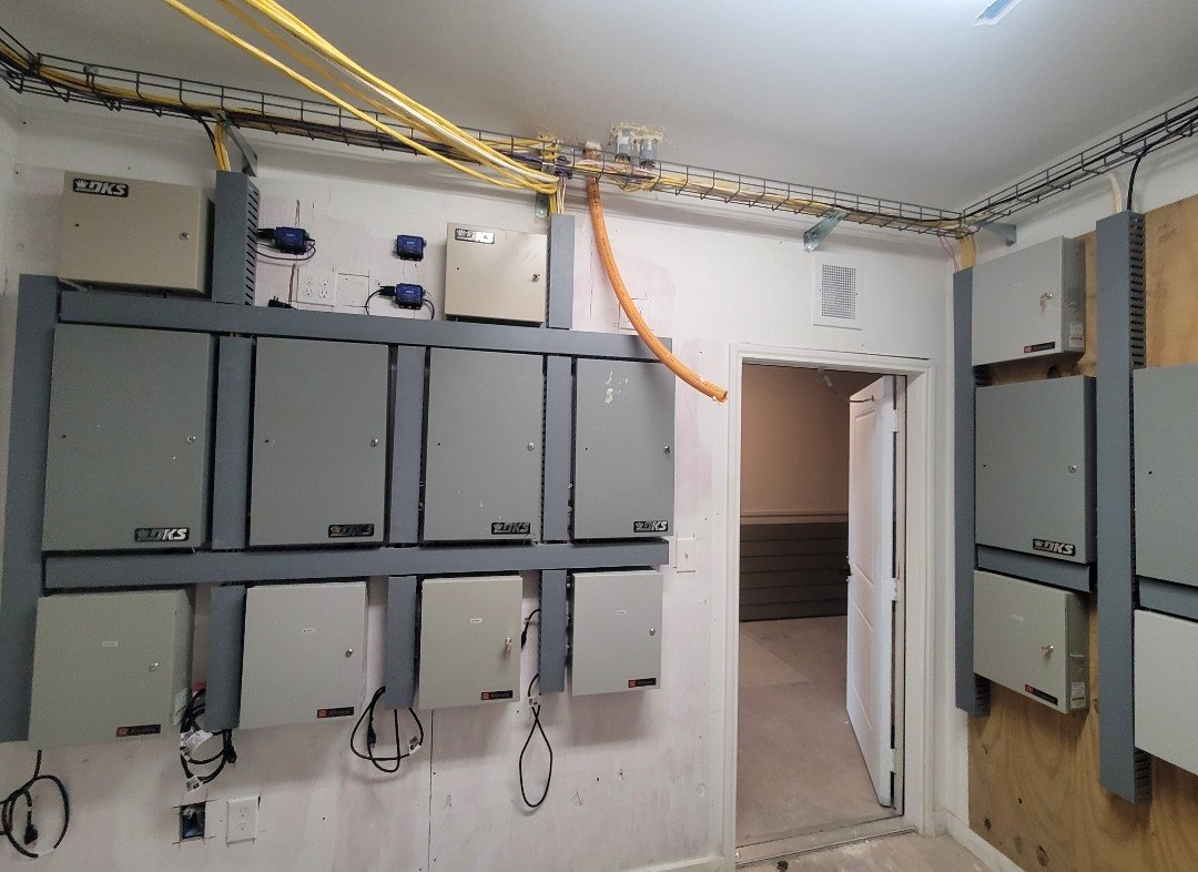 Electrical control panels mounted on the wall in a utility room, with cables running along the ceiling and a doorway leading to another room.