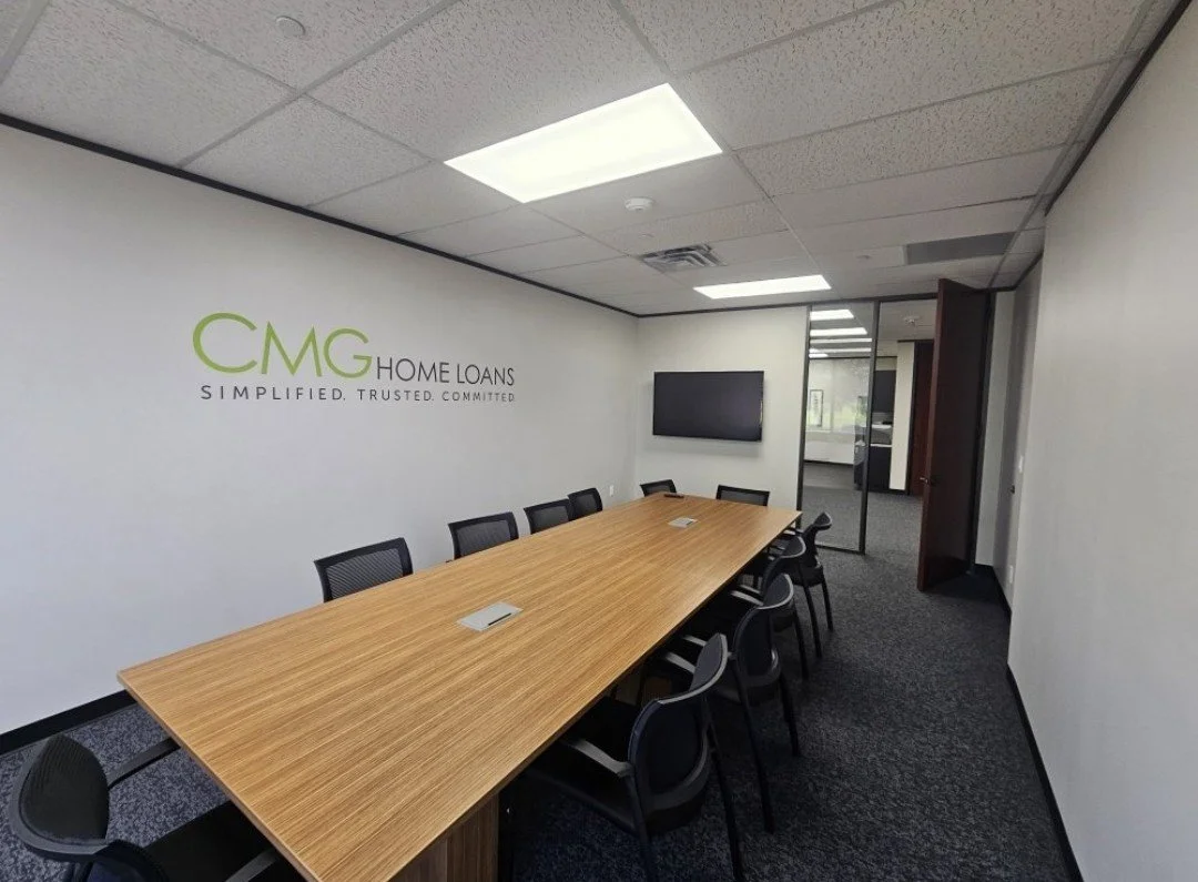 A conference room with a large wooden table surrounded by black chairs, a wall-mounted television, and a logo on the wall that says "CMG HOME LOANS" with the tagline "Simplified. Trusted. Committed."